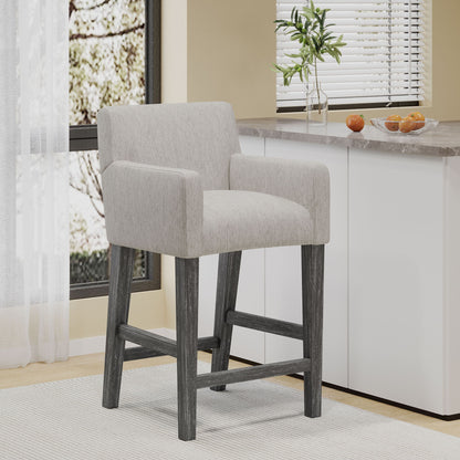 The Charmed Upholstered Counter Stool With H Frame Base And Round Arms, featuring light gray fabric and dark wooden legs, sits in a bright kitchen near a window beside a counter with a plant and oranges.