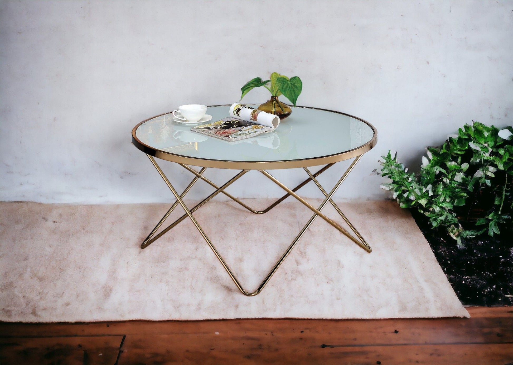 The Valora - Glass Coffee Table with gold metal legs sits on a light rug. On it are a cup and saucer, a rolled-up magazine, and a small plant. Green plants stand nearby against a plain white wall.