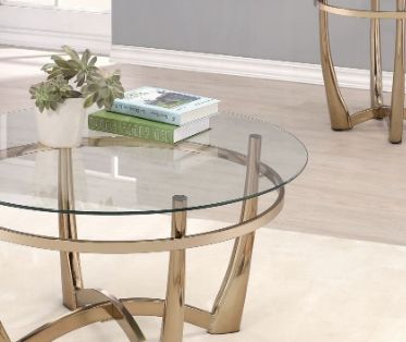 The Orlando - Clear Glass Coffee Table in champagne supports a small potted plant and two books. A matching side table appears in the background, all set on a light wood floor.