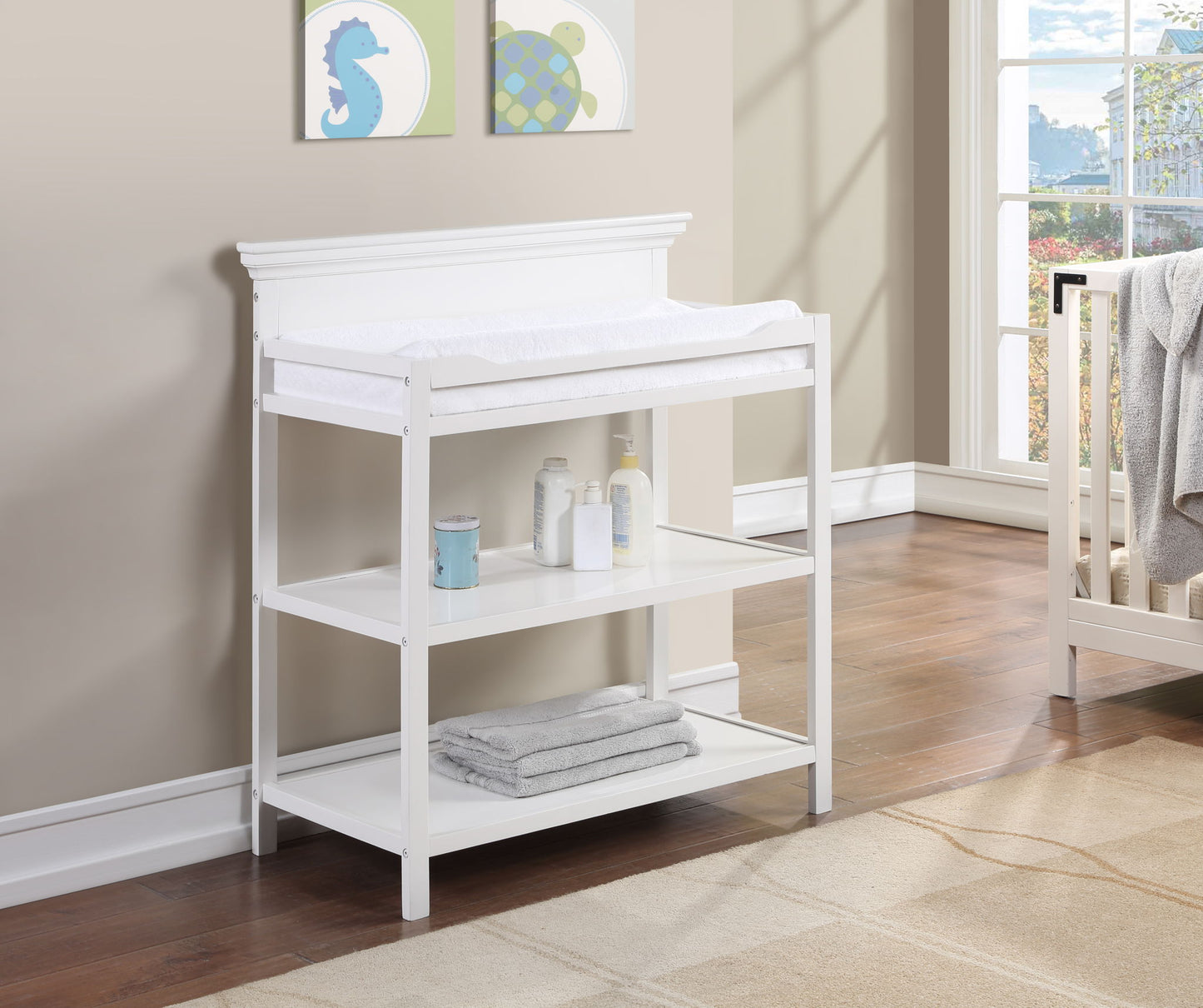 The Universal - Changing Table in white, featuring two shelves for baby essentials and towels, stands by a bright window. Framed sea-themed art decorates the beige wall, while a crib appears on the right.