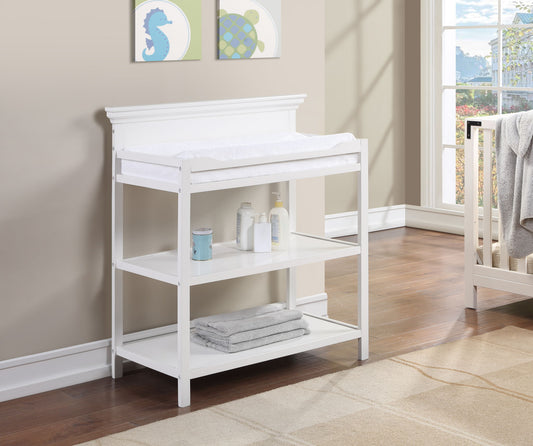 The Universal - Changing Table in white, featuring two shelves for baby essentials and towels, stands by a bright window. Framed sea-themed art decorates the beige wall, while a crib appears on the right.
