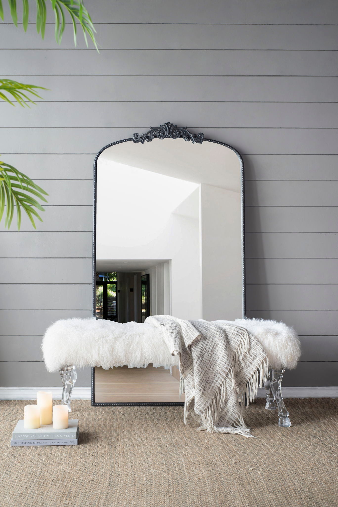 An Arch Mirror, Baroque Inspired Wall Decor, rests against a gray shiplap wall behind a clear acrylic bench with a white throw and plaid blanket. Three pillar candles and magazines are on the floor nearby, with a green plant hanging to the left.