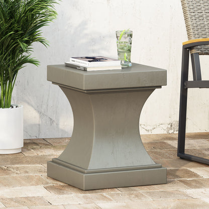 The Athena - Modern Side Table With Pedestal Design, in square gray, sits on a patio with a glass of lime water and two books on top. A potted plant and wicker chair are nearby against a white wall.