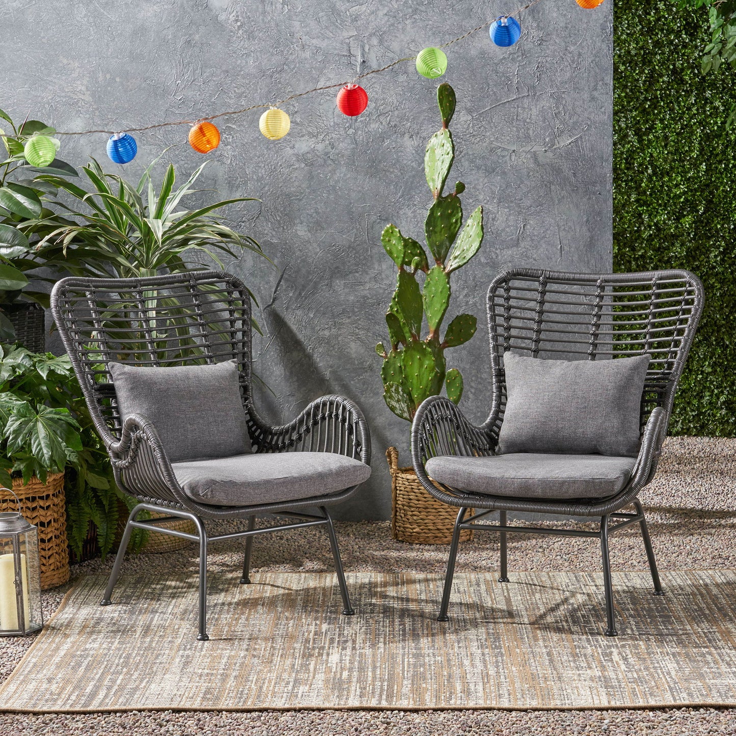 Two Modern Aesthetic Outdoor Rattan Accent Chairs with gray cushions sit on a textured rug outdoors among green plants, while colorful string lights hang above against a gray wall, creating a cozy patio vibe.
