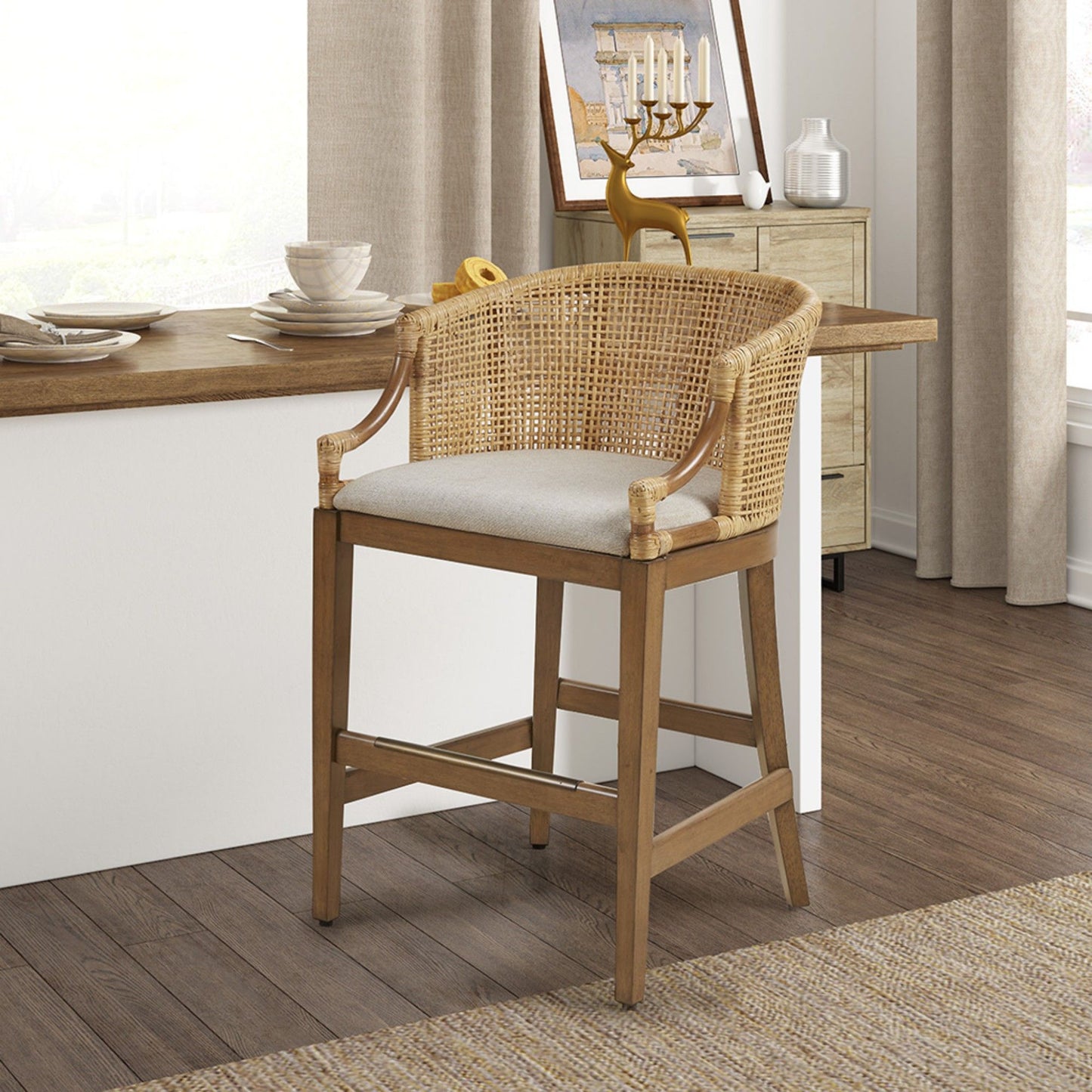 The Casual Handcrafted Rattan Counter Stool, featuring a cane backrest and light cushioned seat, is placed at a kitchen counter in a bright space with wood floors, light curtains, and neutral decor.
