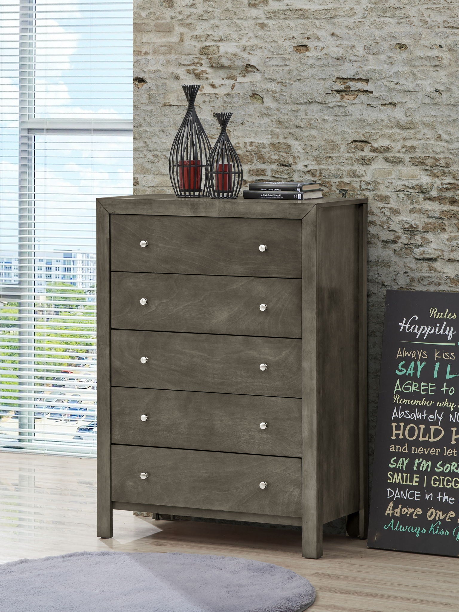 The Modern Elegant Transitional Storage Chest, featuring five gray wooden drawers with silver knobs, stands on a light floor beside a decorative sign and two candle holders. Large windows and a stone wall are seen behind it.