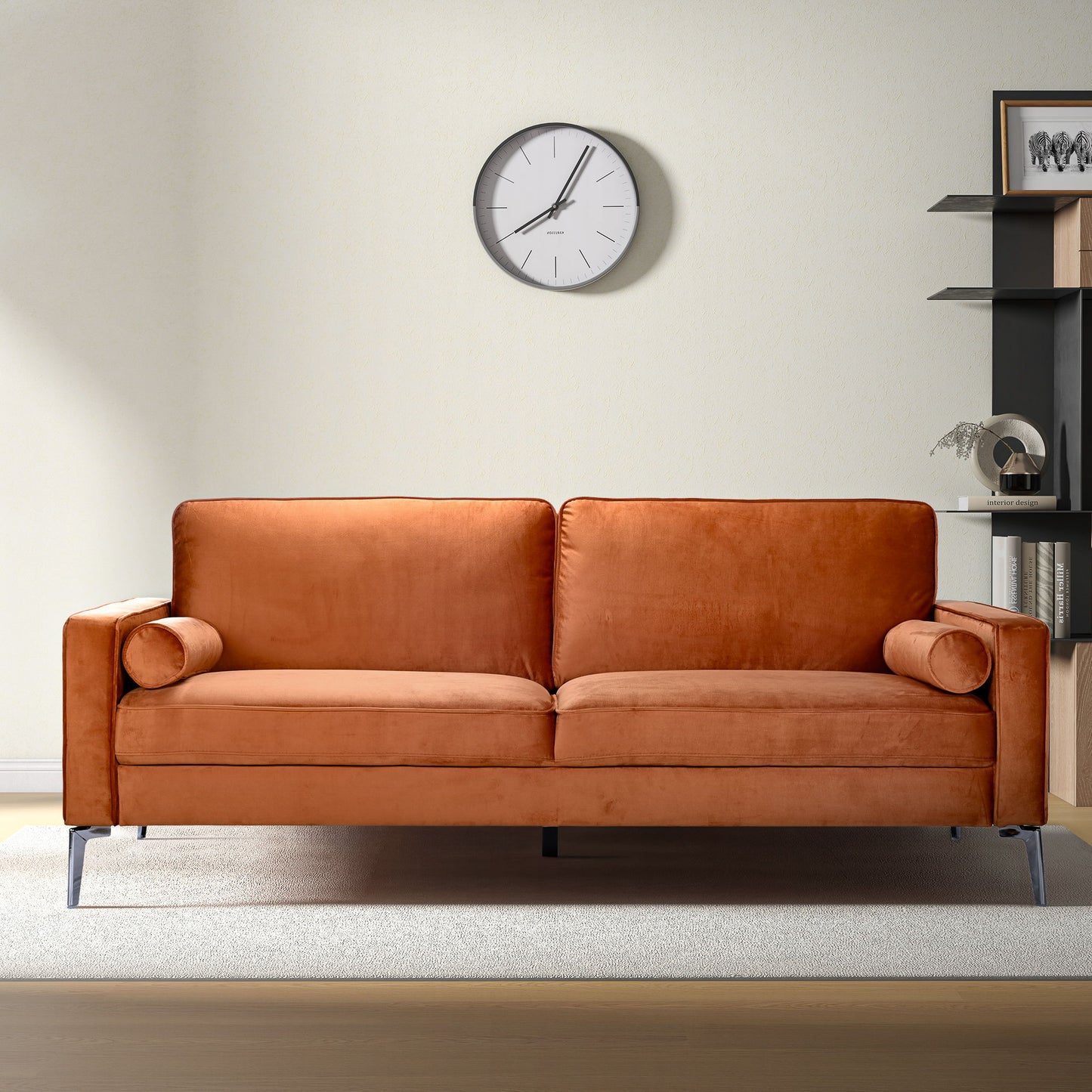 A modern living room featuring the Axten - Sofa - Orange with two matching bolster pillows, a light rug, a wall clock, and black shelves with decor against a cream-colored wall.
