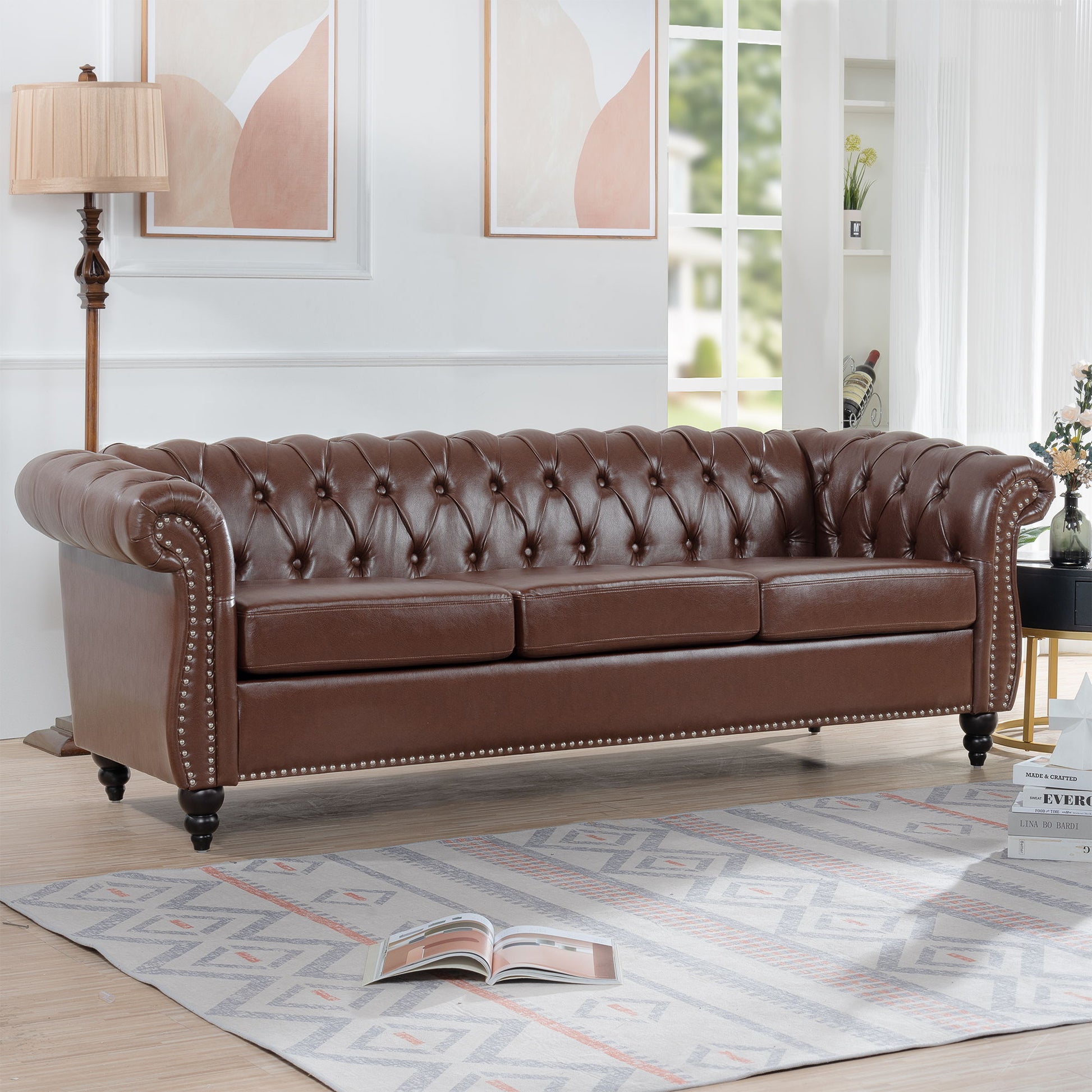 The Rolled Arm Chesterfield 3 Seater Sofa with tufted brown leather and silver studs is featured in a bright living room with light wood floors, a patterned rug, floor lamp, wall art, and an open book on the rug.