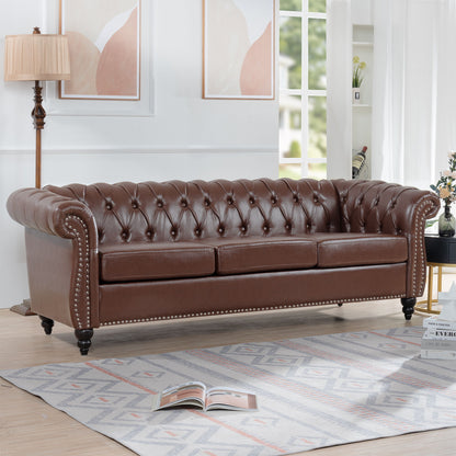 The Rolled Arm Chesterfield 3 Seater Sofa with tufted brown leather and silver studs is featured in a bright living room with light wood floors, a patterned rug, floor lamp, wall art, and an open book on the rug.