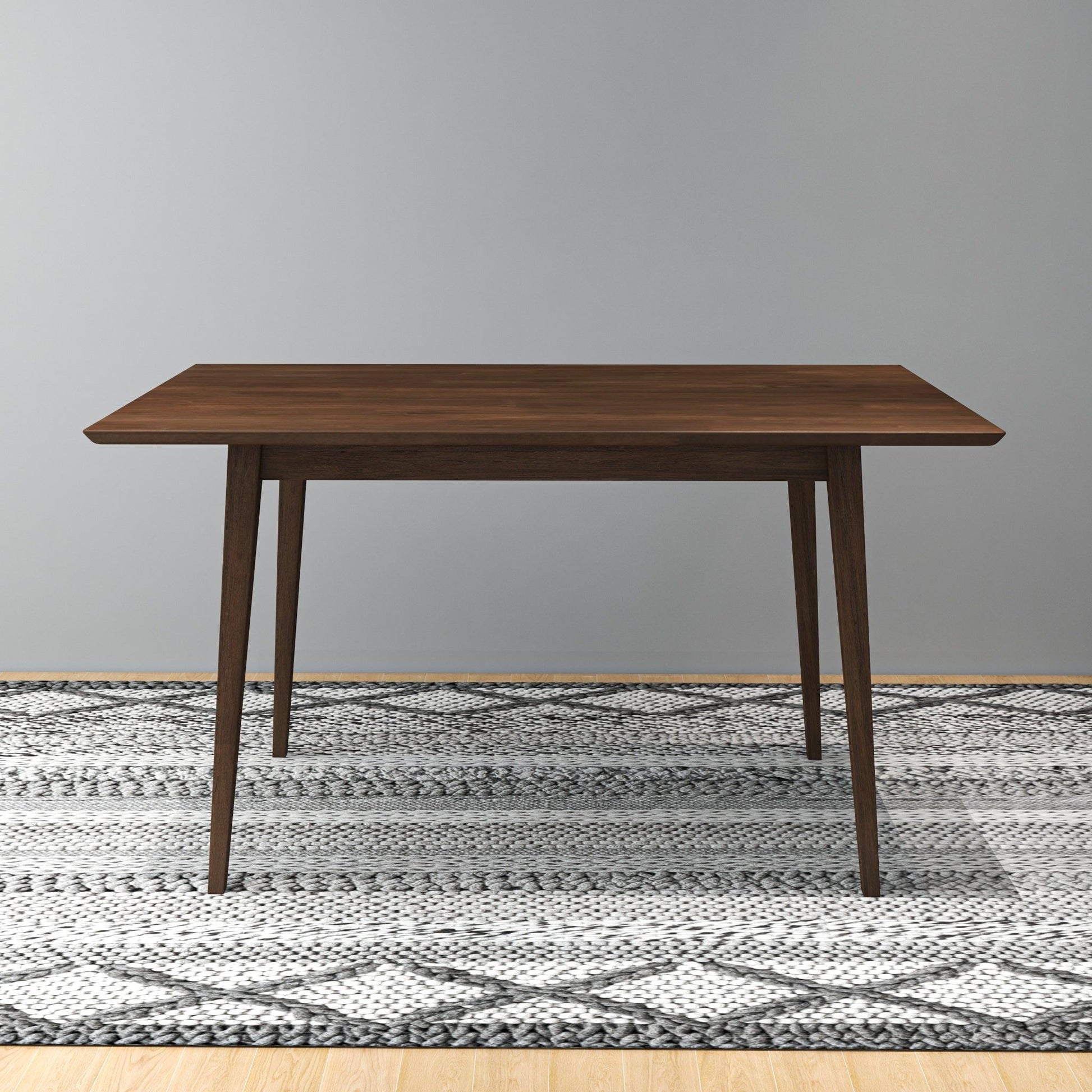The Carlos Dining Table, featuring a rectangular wooden top and tapered legs, stands on a patterned gray and white rug against a plain light gray wall.