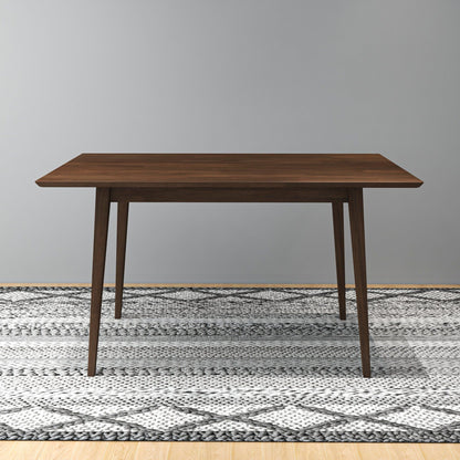 The Carlos Dining Table, featuring a rectangular wooden top and tapered legs, stands on a patterned gray and white rug against a plain light gray wall.