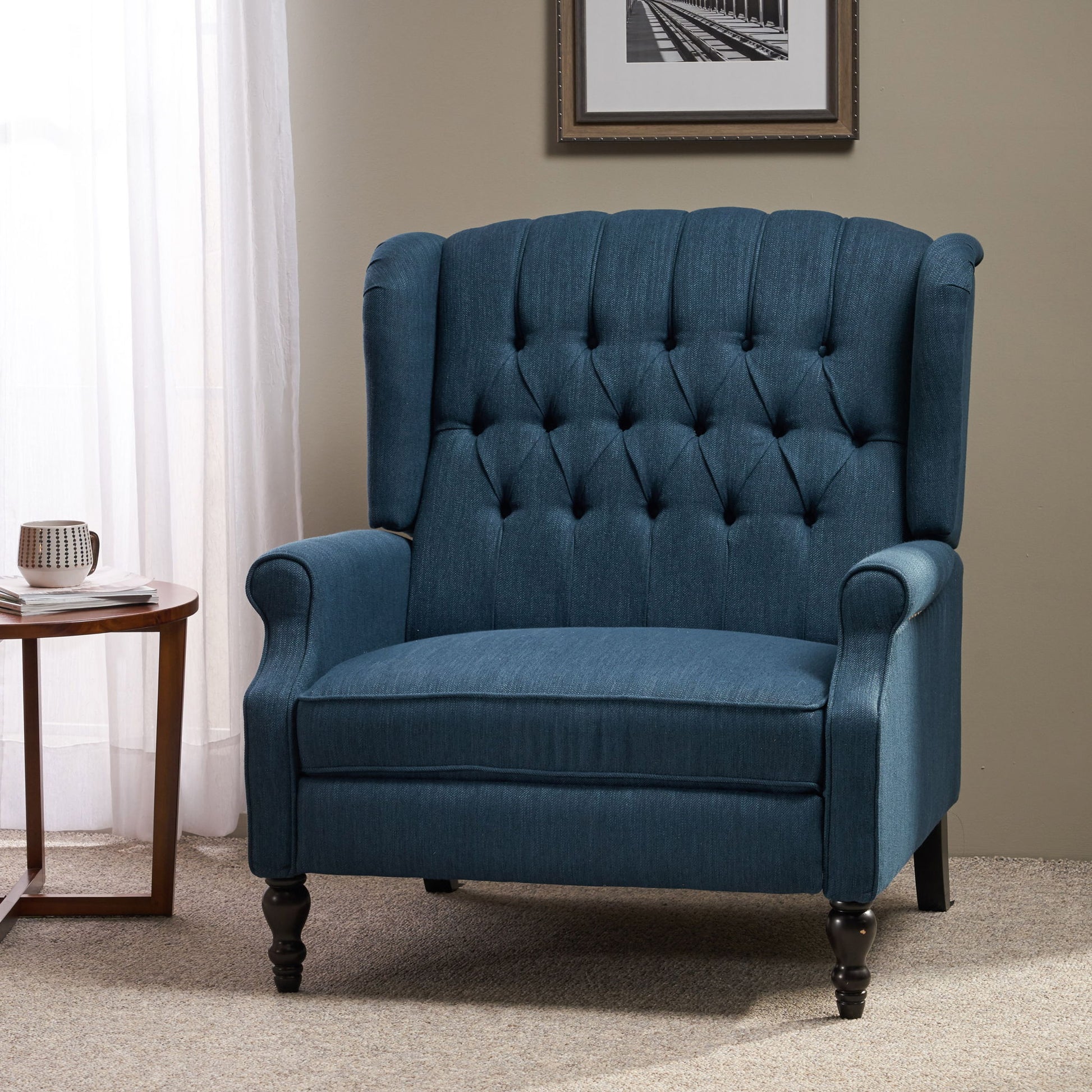 The Dunham Contemporary Fabric Tufted Oversized Pushback Recliner with dark wooden legs is placed on a beige carpet beside a round wooden side table holding a mug and book, near a window with white curtains and a framed photo.