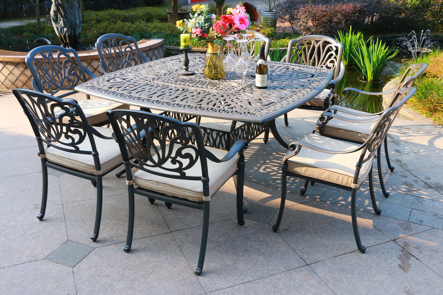 The Square 8 Person 64" Long Aluminum Dining Set With Cushions, arranged on a stone patio by a pond and greenery, features cream-cushioned chairs and a spacious table set with glassware, wine, and flower vases.
