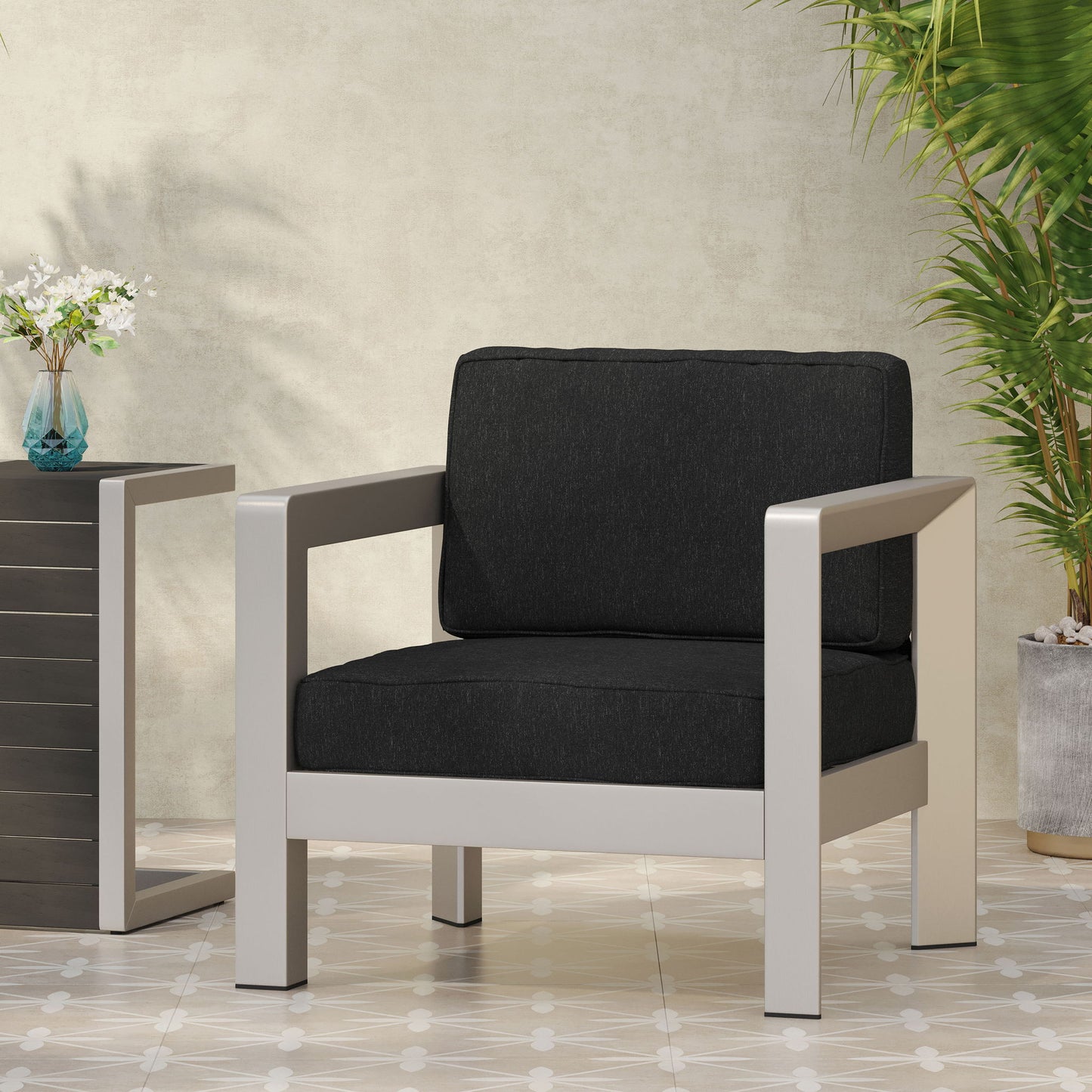 The Aviara - Luxurious Outdoor Club Chair with black cushions and a light metal frame sits on a tiled floor beside a sleek side table, surrounded by green plants.
