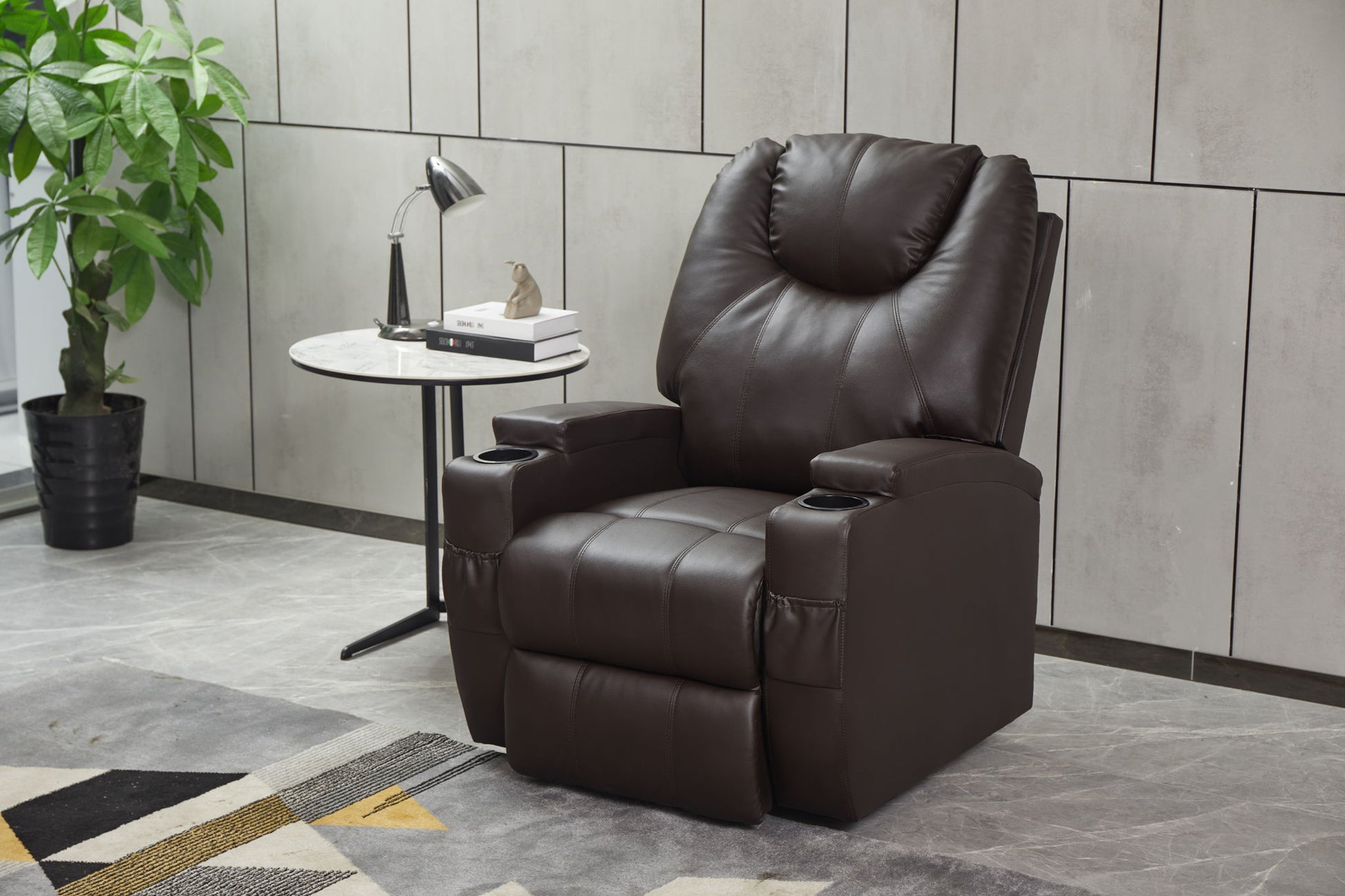 A Modern Recliner Chair With Cup Holders and Remote Control sits beside a round marble side table holding a lamp, books, and decor. A potted plant and part of a patterned rug are visible in the stylish room.