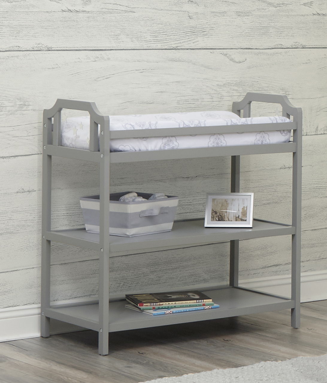 The Celeste - Changing Table in gray wood has a padded top, a storage bin on the middle shelf, and children's books on the bottom shelf. It’s displayed against a light wood-paneled wall and set on a wood floor.