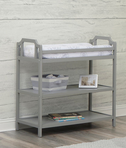 The Celeste - Changing Table in gray wood has a padded top, a storage bin on the middle shelf, and children's books on the bottom shelf. It’s displayed against a light wood-paneled wall and set on a wood floor.