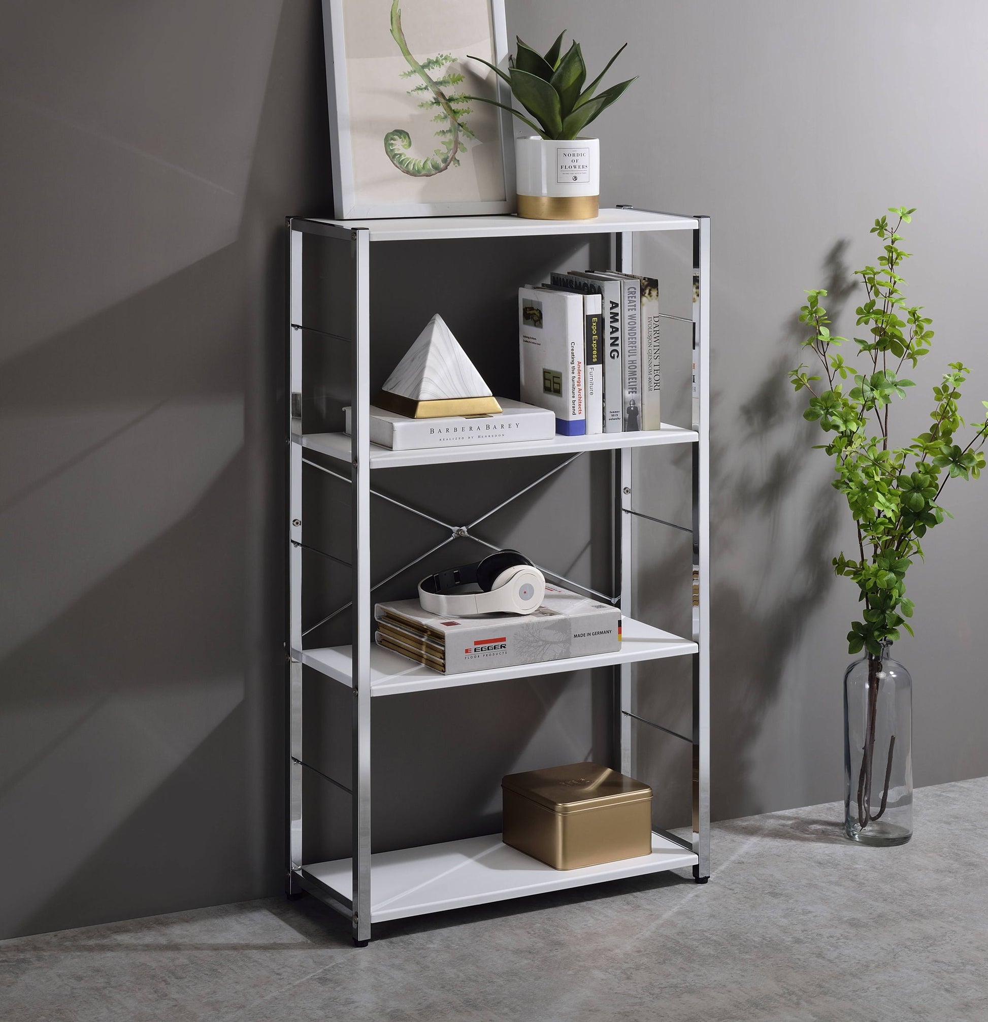 The Tennos - Bookshelf - White features four shelves for books, decor, a potted plant, and headphones. On top sit a framed artwork and plant, while a vase with green branches stands nearby against the gray wall.
