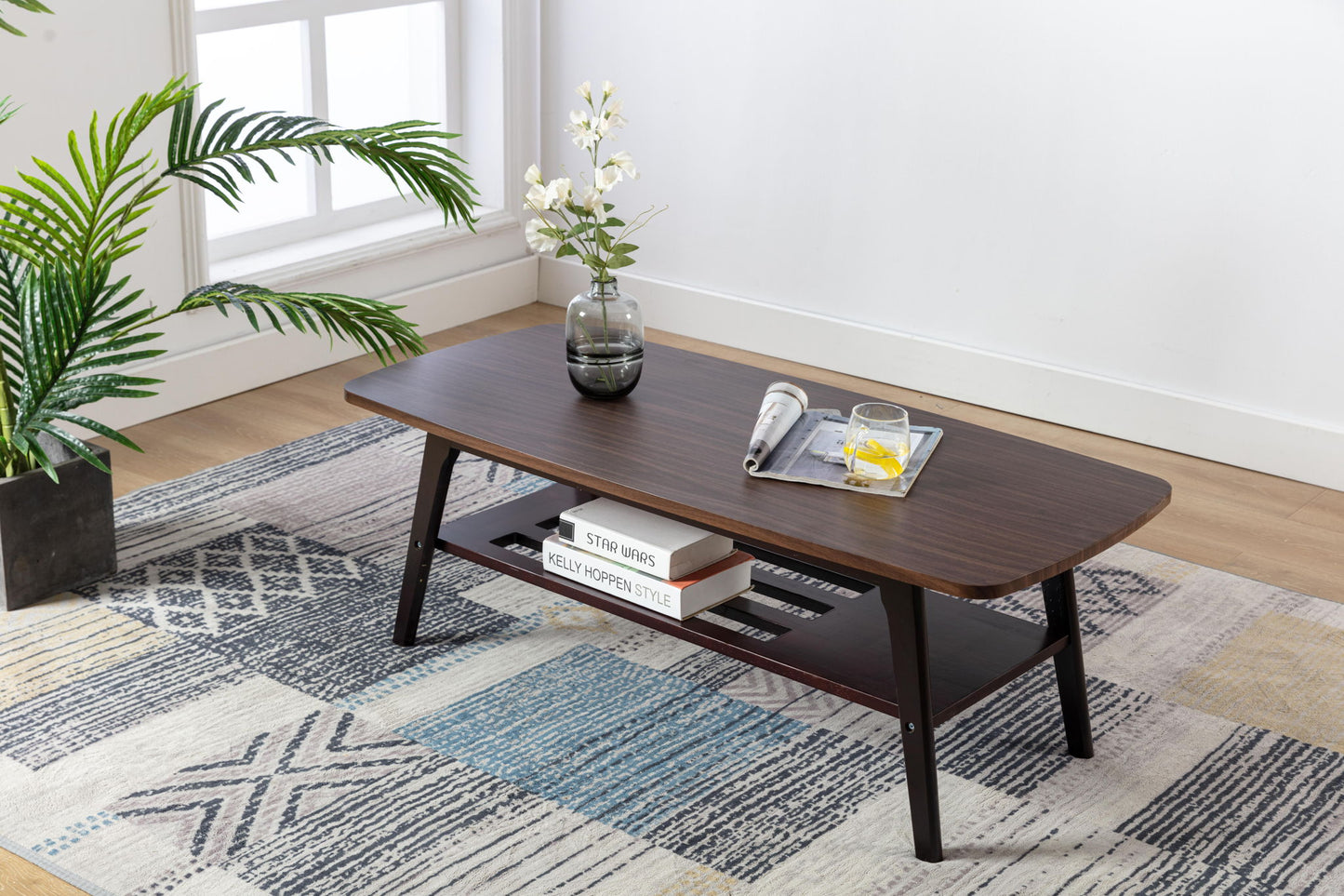 The Bahamas - Coffee Table holds a glass vase with flowers, a magazine, and a drink on top. Books are stored on its lower shelf. The table sits on a patterned rug by the window next to a potted plant.