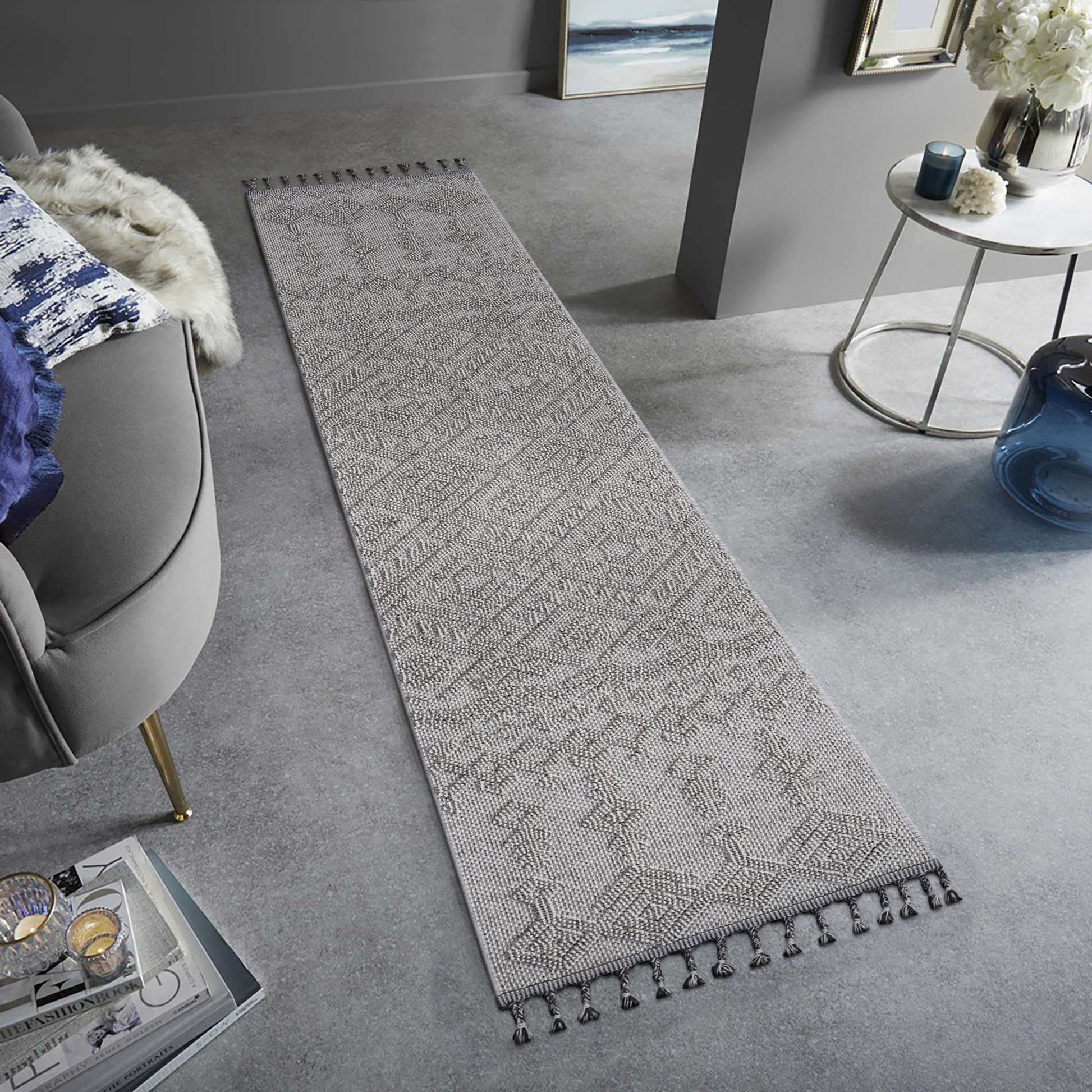 The Guros - Traditional Indoor / Outdoor Area Rug, featuring a long, narrow gray pattern with tasseled edges, is styled on a light gray floor in a modern living room with neutral decor and a gray chair.