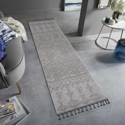 The Guros - Traditional Indoor / Outdoor Area Rug, featuring a long, narrow gray pattern with tasseled edges, is styled on a light gray floor in a modern living room with neutral decor and a gray chair.