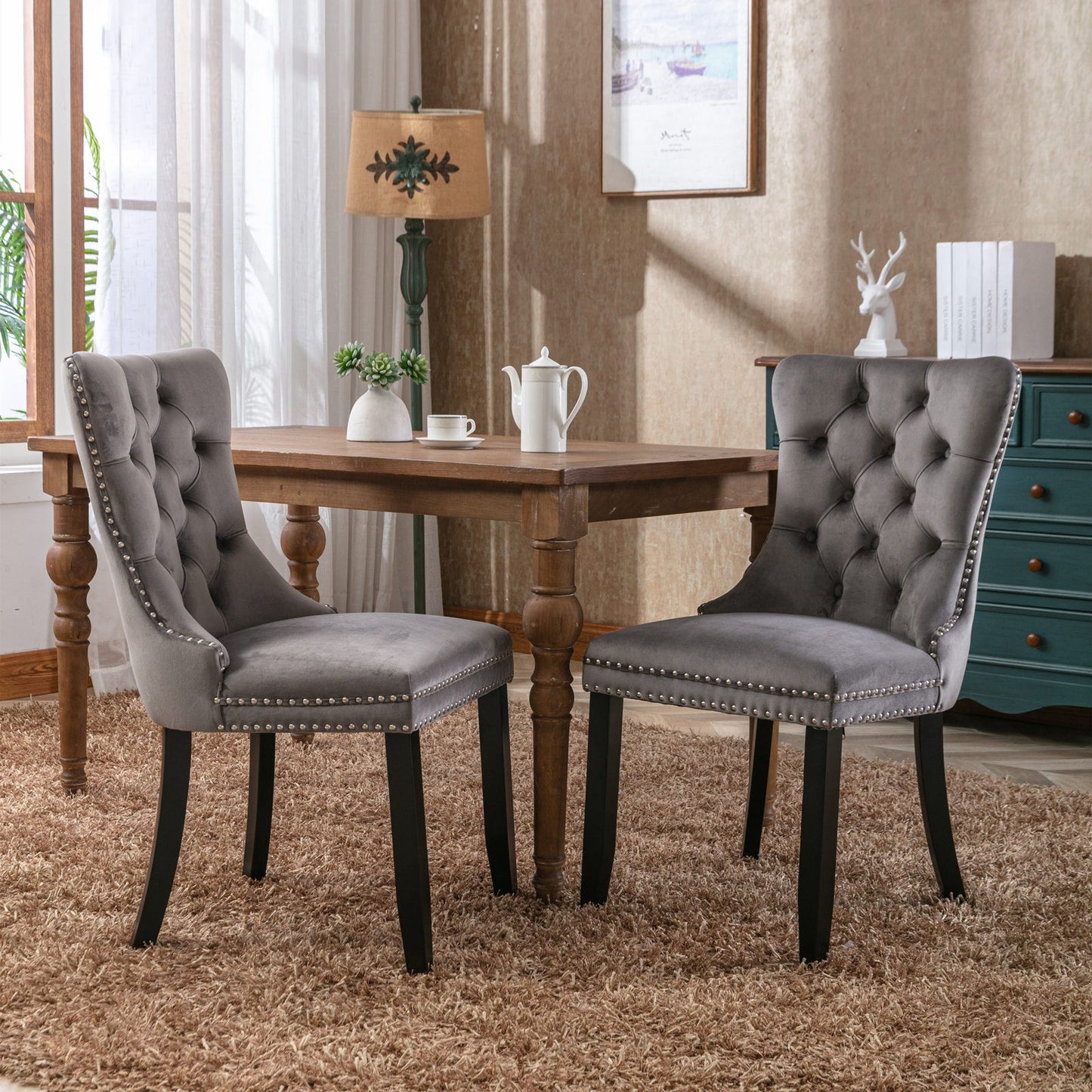 Two Nikki Modern Velvet Upholstered Dining Chairs with tufted details, wood legs, and nailhead trim are set around a wooden table in a bright room featuring a shaggy carpet, lamp, potted plant, and drawers.