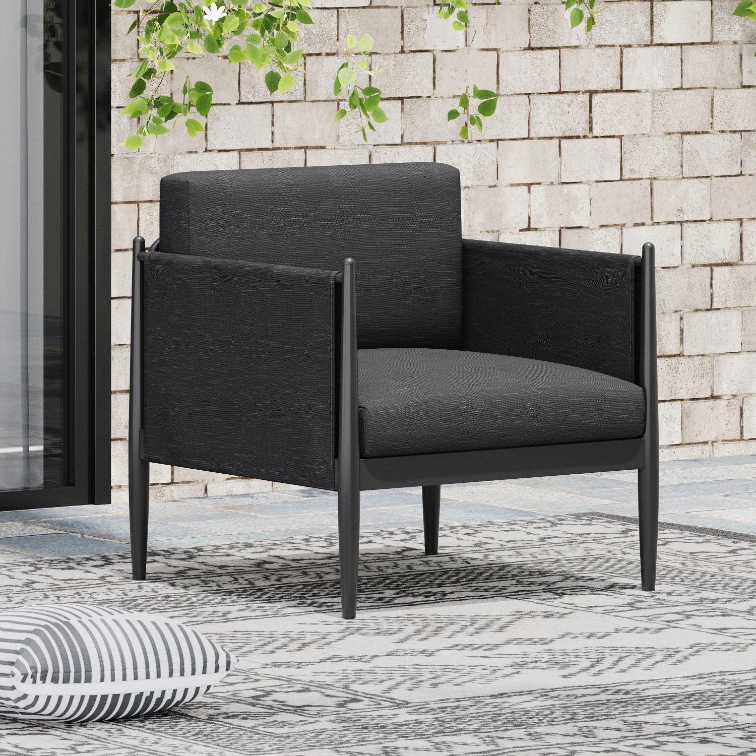 The Dawson - Club Chair, a modern black armchair with clean lines, is set on a patterned outdoor rug by a textured white brick wall and greenery for a stylish patio look. A striped pillow rests on the ground nearby.