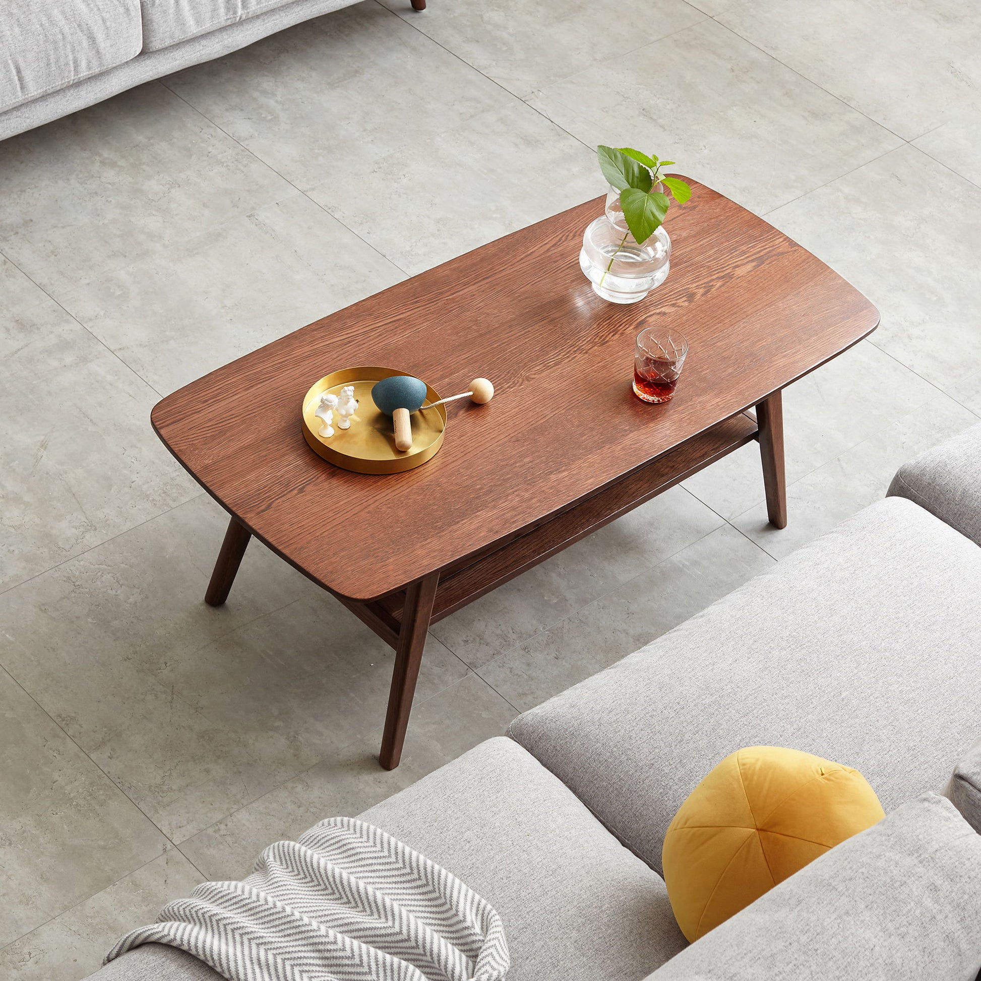 A Low Coffee Table With Storage Shelf holds a glass vase with a green plant, a gold tray with decor items, and a glass of red drink, surrounded by light gray sofas and a yellow cushion on tiled flooring.
