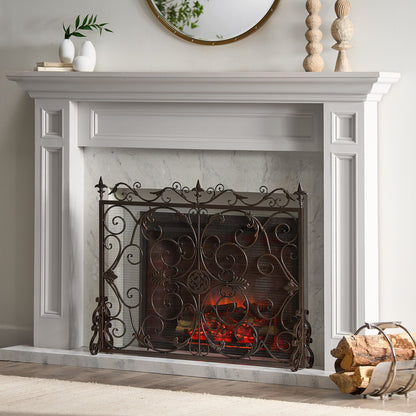 A white fireplace features the Darcie - Wrought Fireplace Screen with an ornate design, glowing embers, a log holder of firewood, and simple decor such as a round mirror, vases, and a wooden beaded ornament on the mantel.