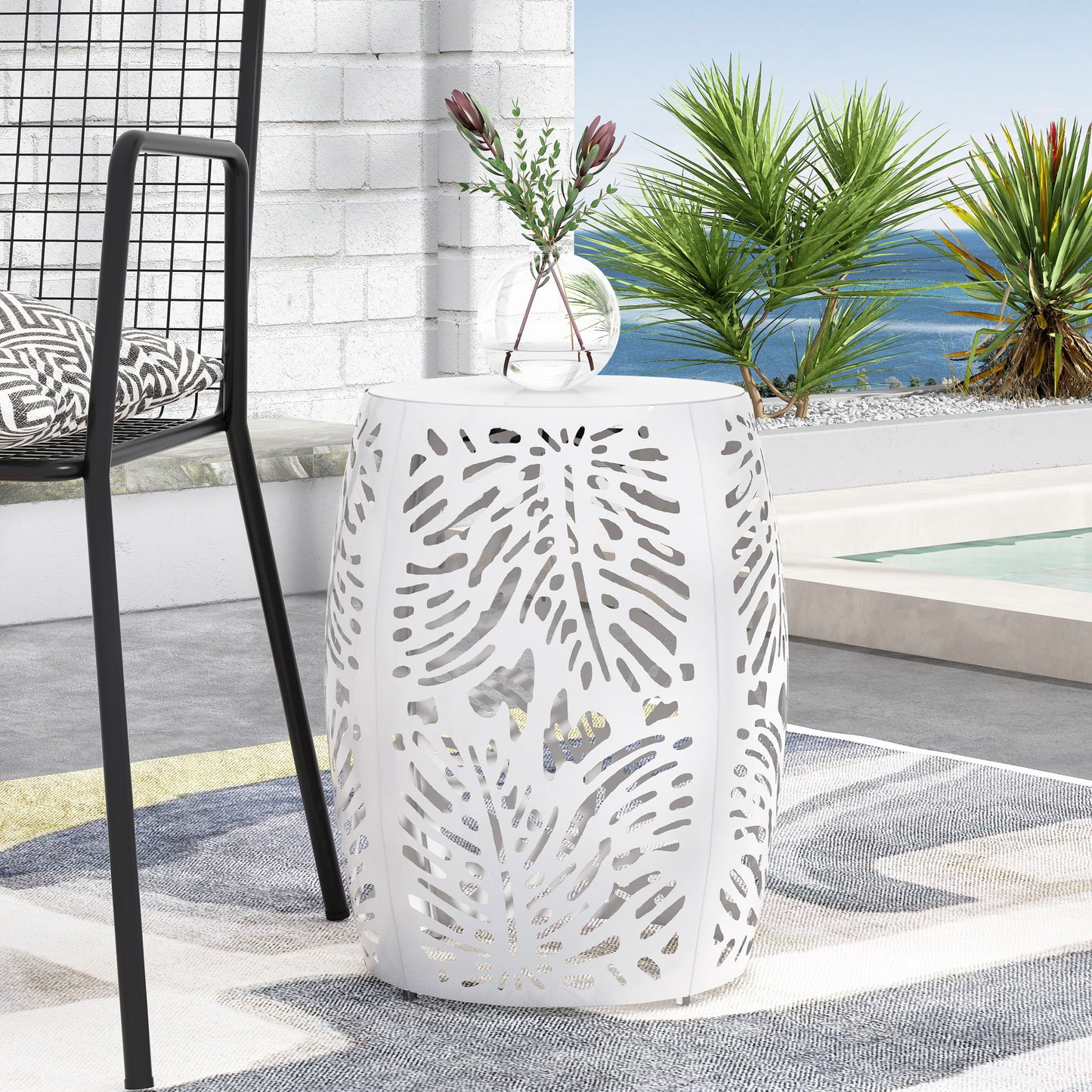 A Modern Design Metal End Table, Large, featuring cutout leaf patterns, stands on a patterned rug near a pool, with a chair and decorative vase with flowers nearby; palm trees and ocean are visible in the background.