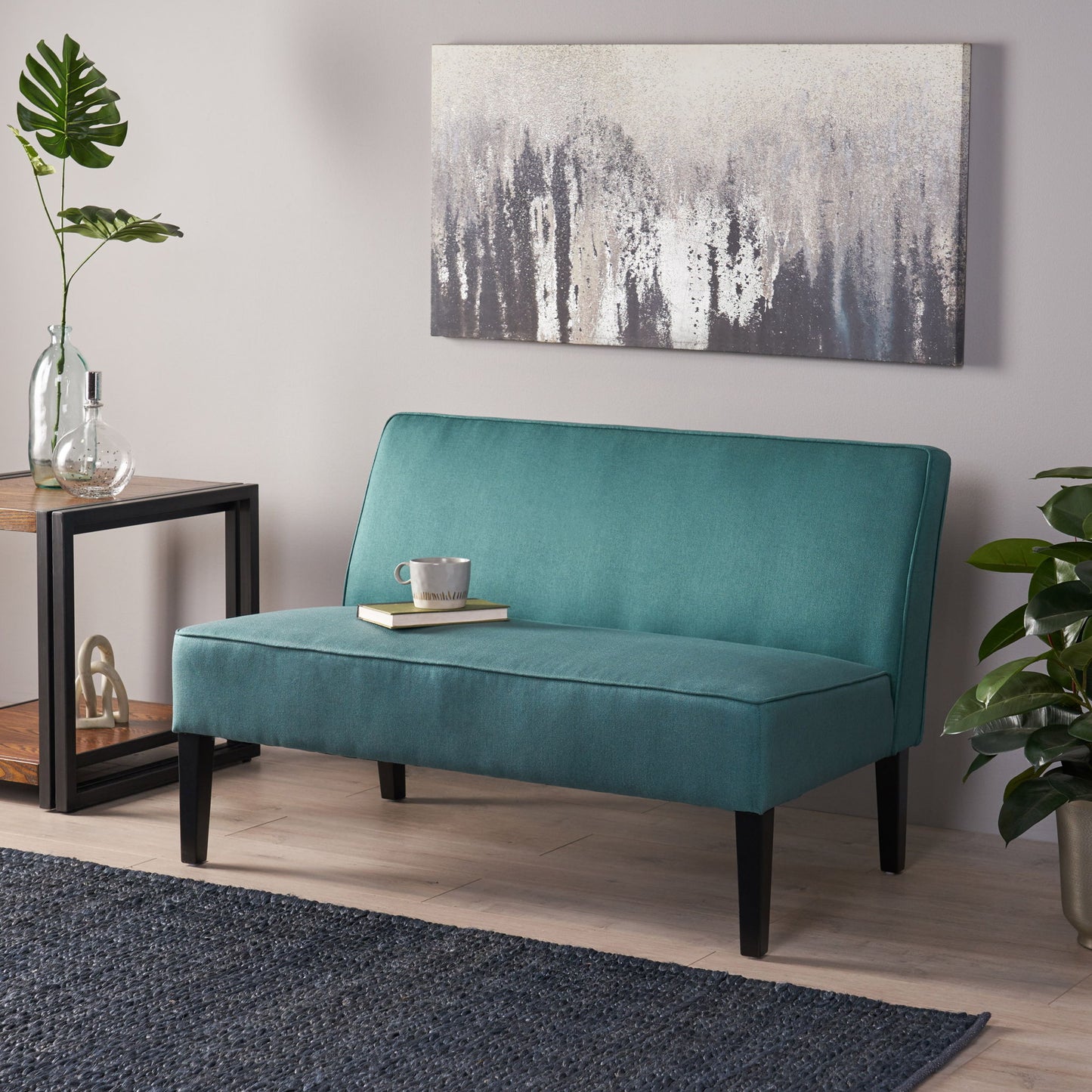 A modern living room featuring a teal Contemporary Loveseat With Upholstered Fabric And Birch Wood Legs, a coffee cup and book on the seat, abstract wall art above, dark rug, side table with plants and decor, and a large leafy plant on the right.