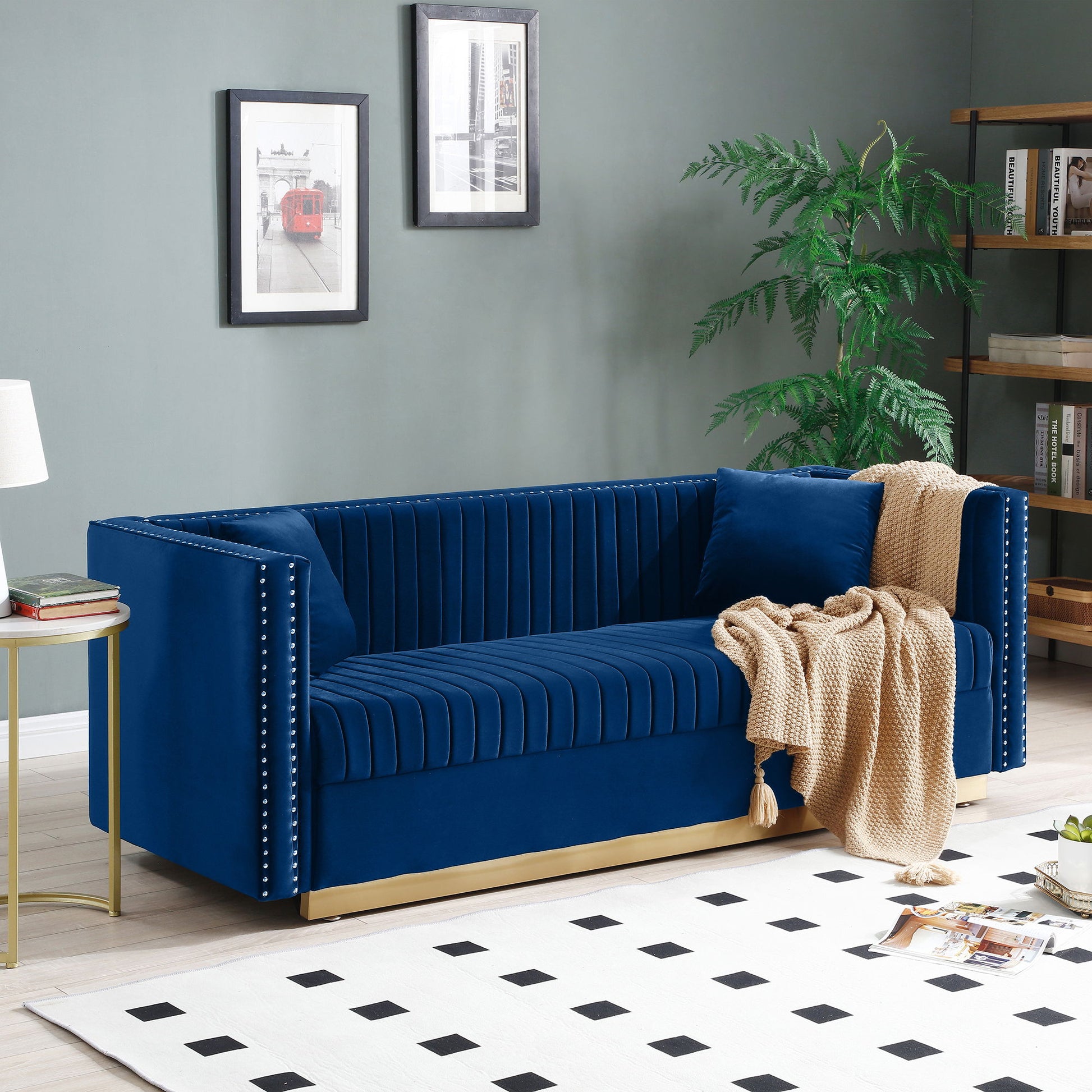 A modern living room with a Contemporary Vertical Channel Tufted Sofa, upholstered in blue velvet with silver studs and two pillows, draped with a beige knit blanket, set against a green wall with framed art and a black-and-white rug.