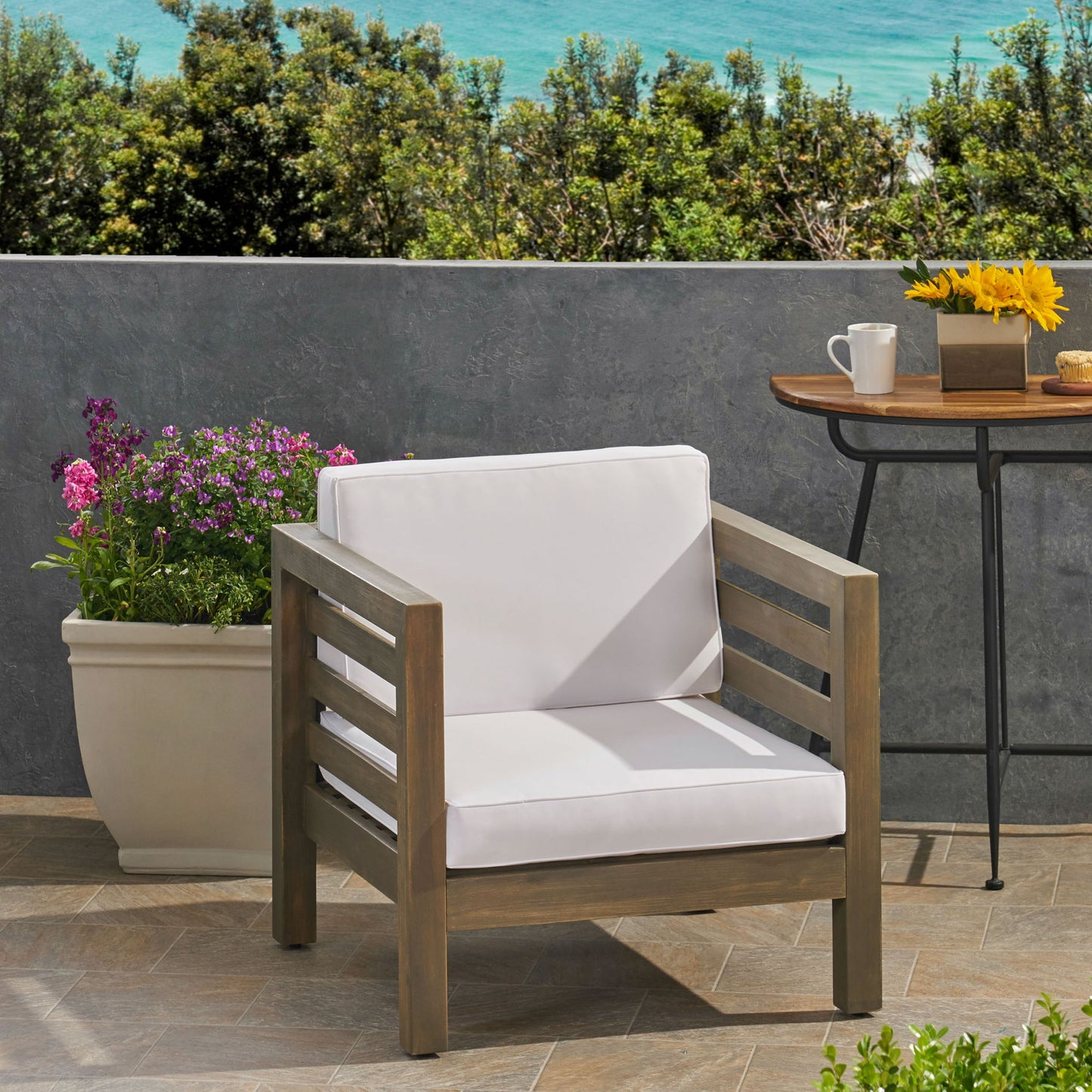 An Outdoor Acacia Wood Patio Chair With Fabric Cushion sits on a tiled patio beside a planter with pink flowers, while a table with sunflowers and views of trees and the ocean are visible in the background.