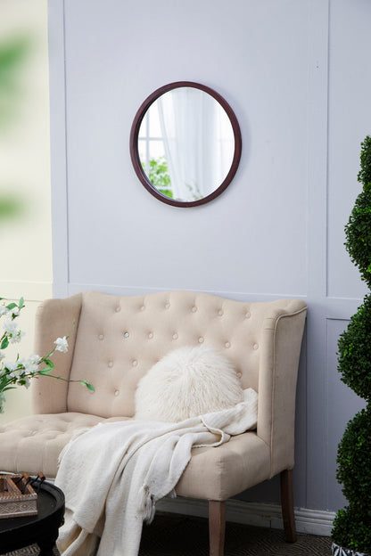 A beige tufted loveseat with a white fluffy pillow and blanket sits against a light gray wall. Above is a Circle Wall Mirror With Wooden Frame for living rooms, reflecting nearby greenery and the room’s window.