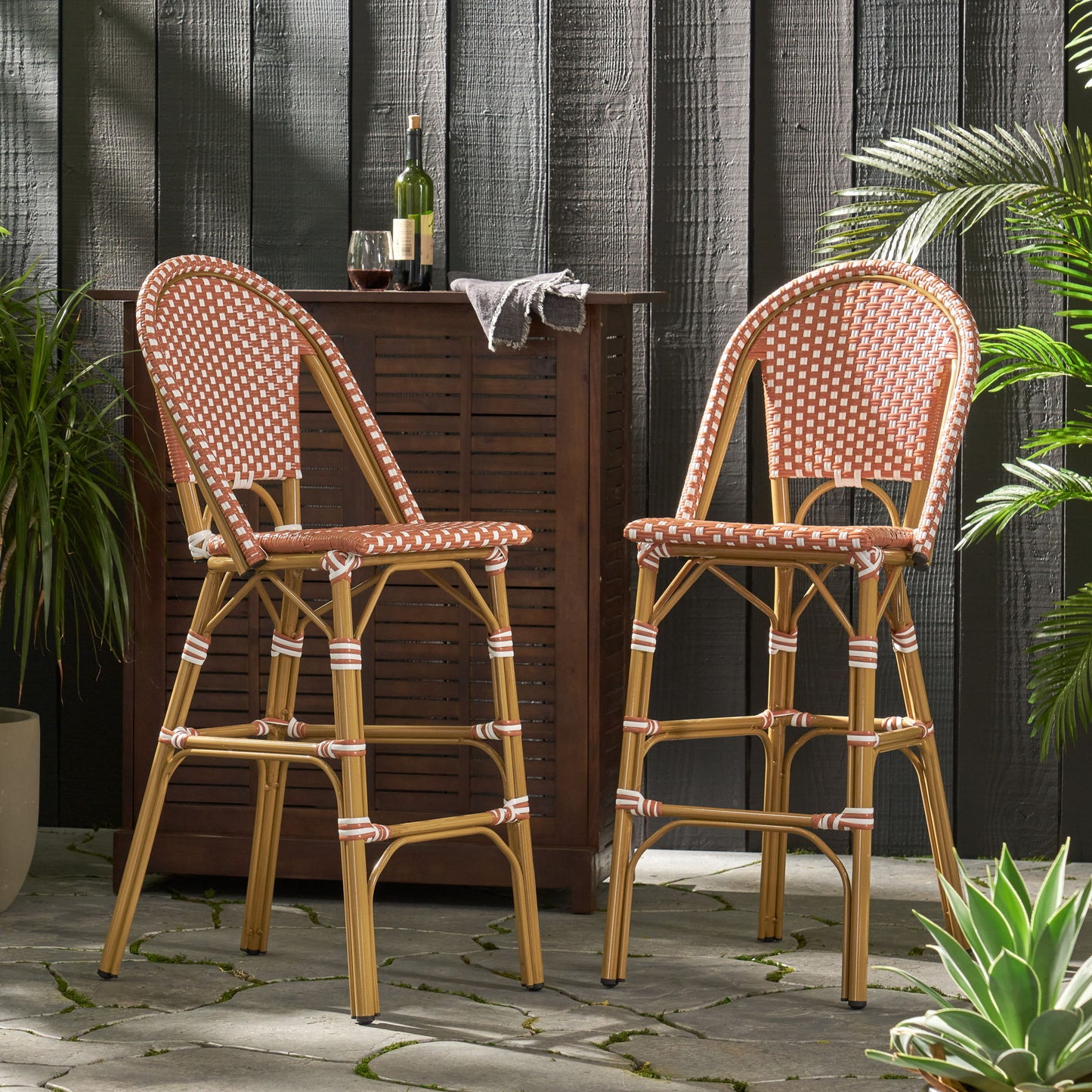 Two Outdoor Pe Rattan French Barstools with red-and-white woven seats stand before a dark wooden outdoor bar, surrounded by wine, glasses, and plants on a stone patio.