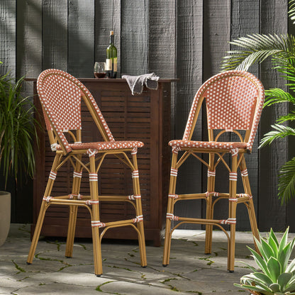 Two Outdoor Pe Rattan French Barstools with red-and-white woven seats stand before a dark wooden outdoor bar, surrounded by wine, glasses, and plants on a stone patio.
