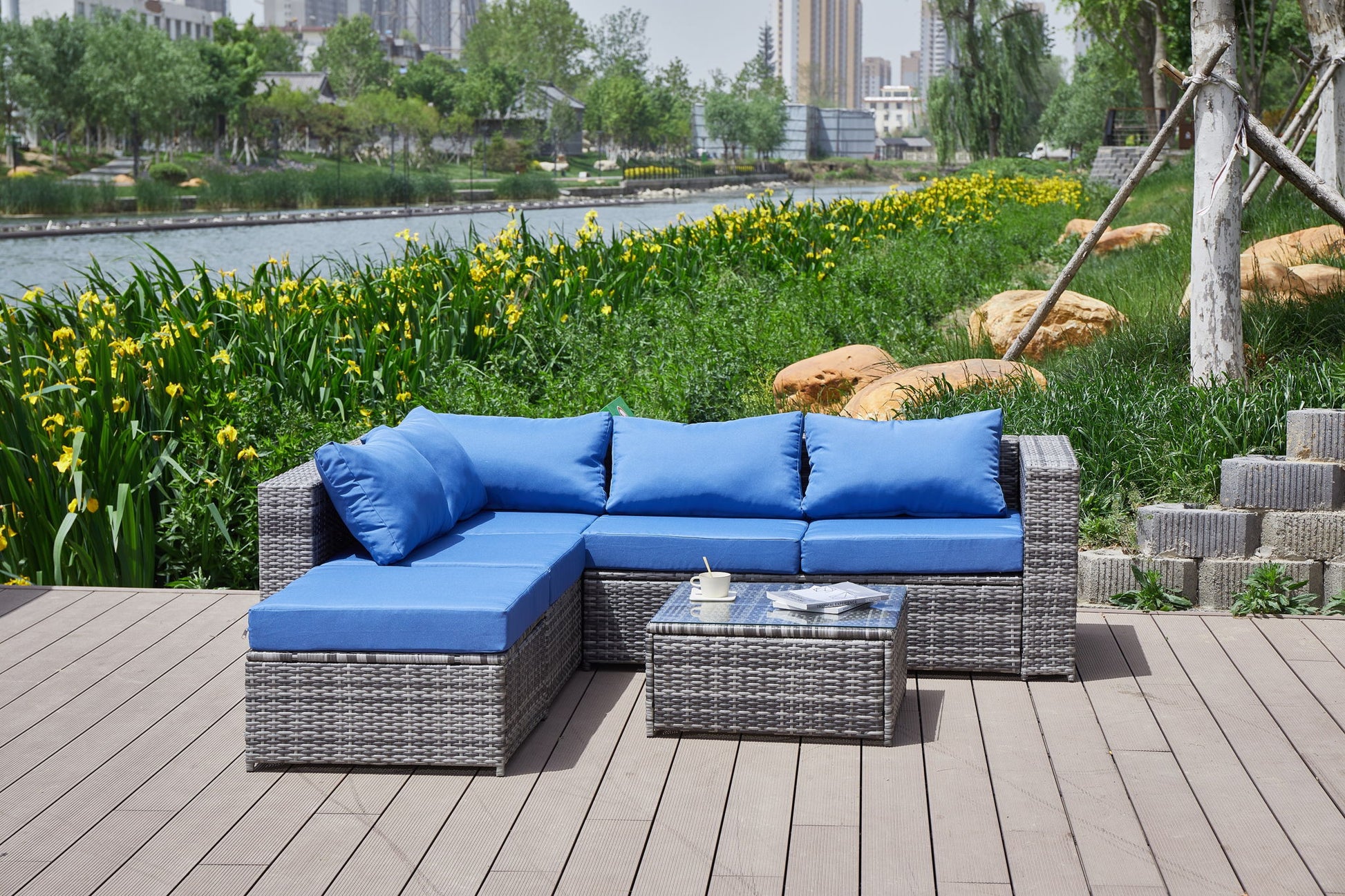 A Patio Sofa With Cushions and a matching coffee table are arranged on a wooden deck by the river, surrounded by greenery and yellow flowers, with city buildings in the background.