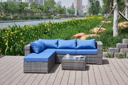 A Patio Sofa With Cushions and a matching coffee table are arranged on a wooden deck by the river, surrounded by greenery and yellow flowers, with city buildings in the background.