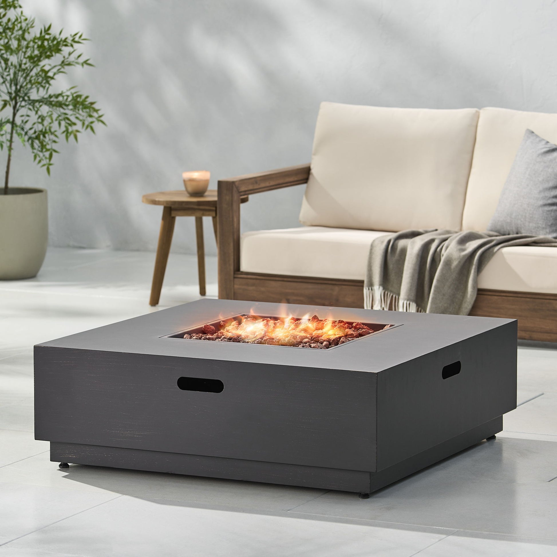A modern patio features a Modern Square Fire Pit With 50,000 Btu glowing with flames. Behind it sits a wooden outdoor sofa with cream cushions, a gray blanket, and a side table topped with a candle and potted plant.