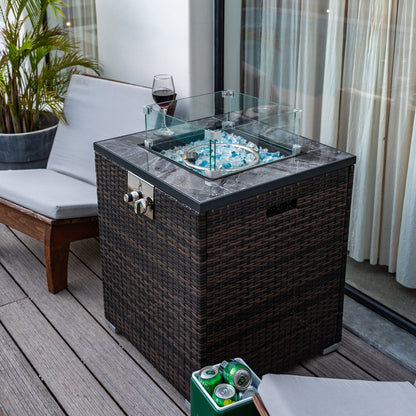A Propane Gas Square Fire Pit with tile tabletop and wicker base features a glass wind guard, blue fire glass, and a wine glass on top. Nearby are a cushioned bench, plant, and drink cans on a wooden patio deck.