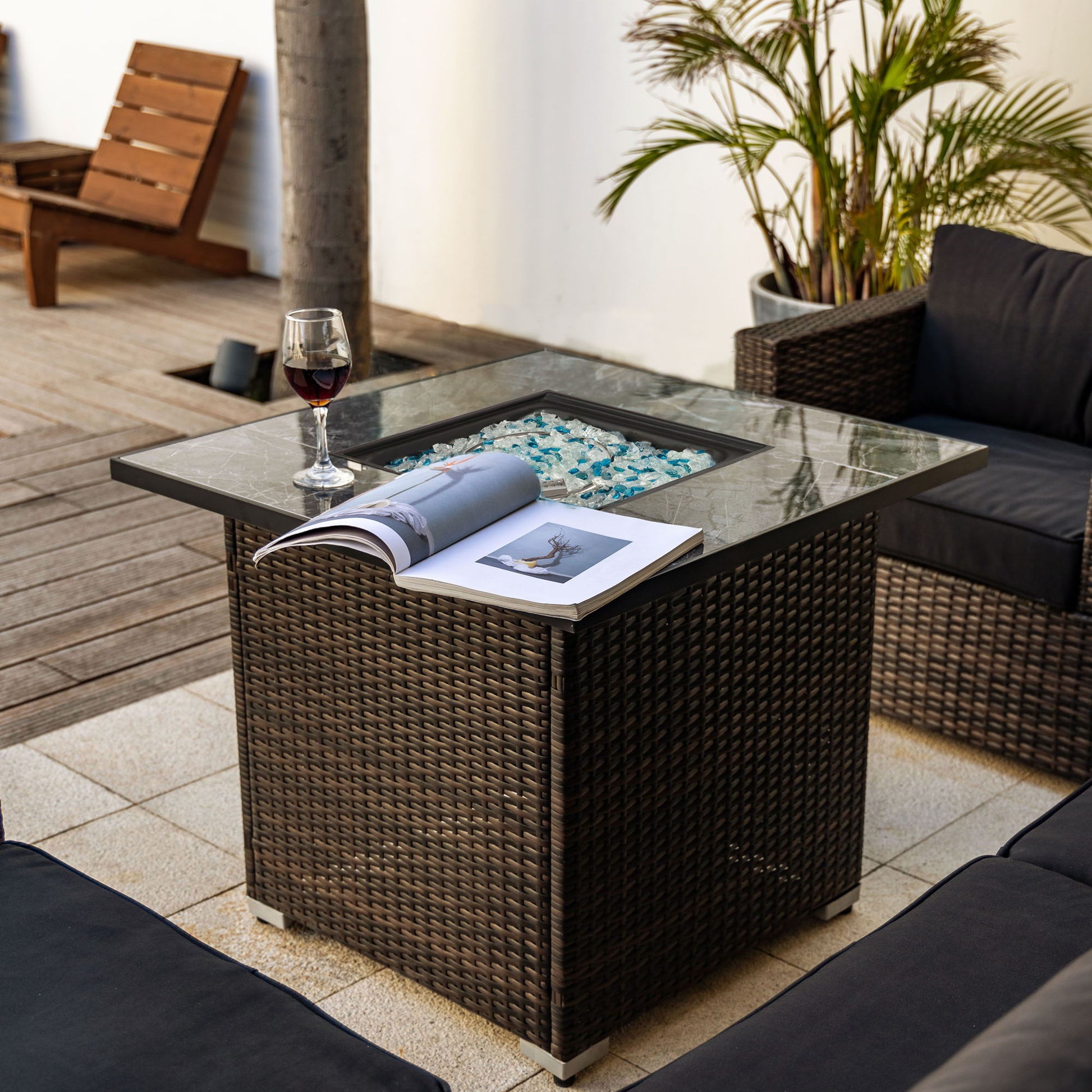 A glass of red wine and an open magazine sit atop the 30" Outdoor Fire Table Propane Gas Fire Pit Table with blue glass rocks, surrounded by outdoor seating, plants, and wooden flooring.