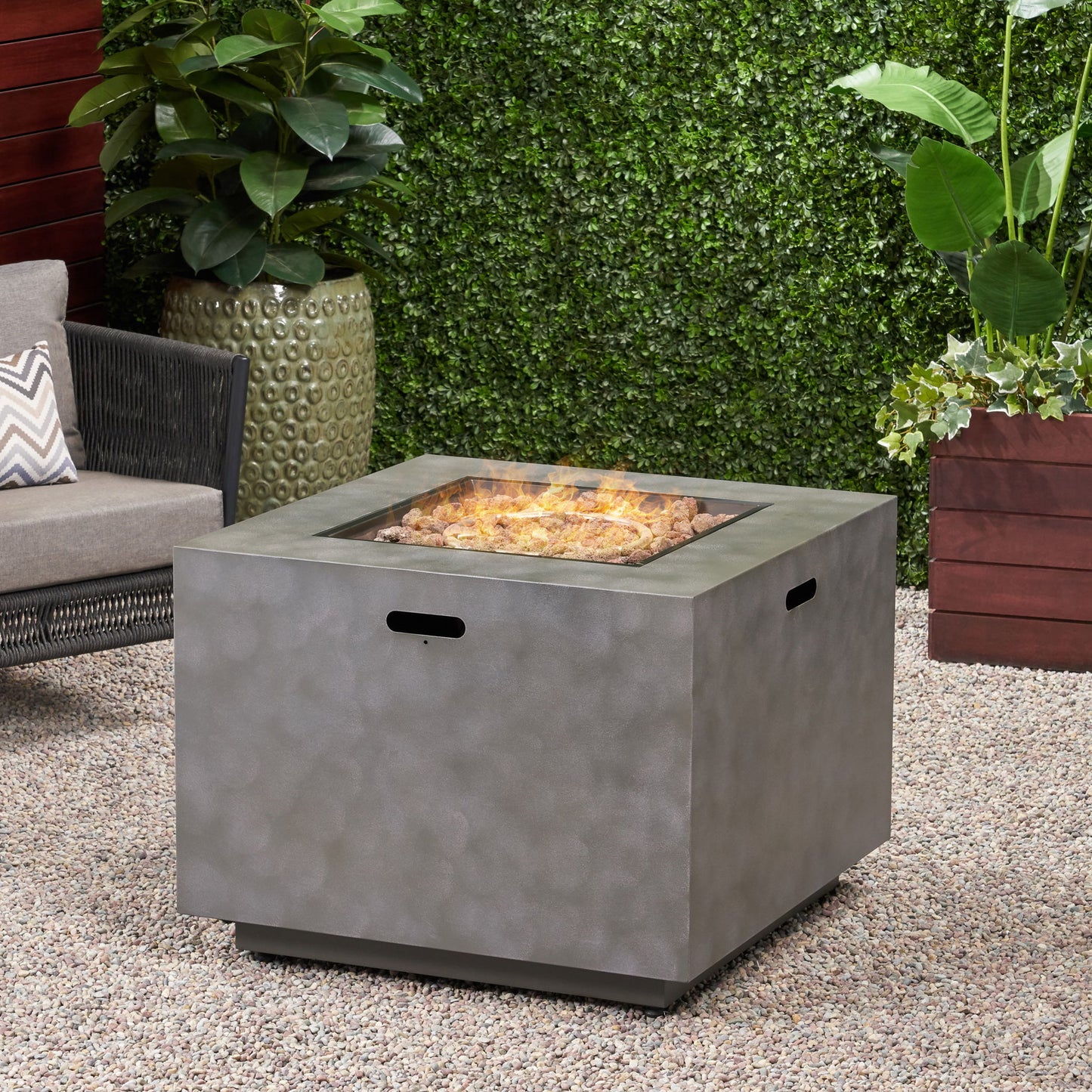 The Outdoor Patio Gas Burning Fire Pit Table 50,000 Btu, Square, with a gray textured finish, sits on a gravel patio among green plants, a wicker chair, and planters near a leafy hedge. Flames flicker in the fire pit.