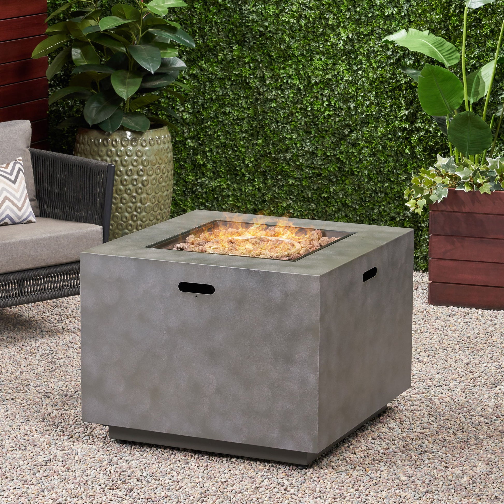 The Outdoor Patio Gas Burning Fire Pit Table 50,000 Btu, Square, with a gray textured finish, sits on a gravel patio among green plants, a wicker chair, and planters near a leafy hedge. Flames flicker in the fire pit.
