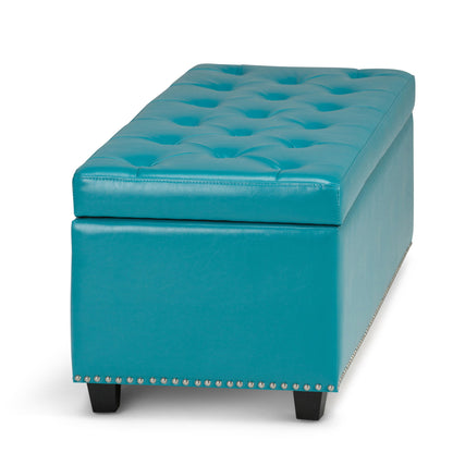 The Hamilton Upholstered Storage Ottoman features a turquoise, rectangular design with tufted top, silver nailhead trim along the base, and short black legs, displayed against a white background.