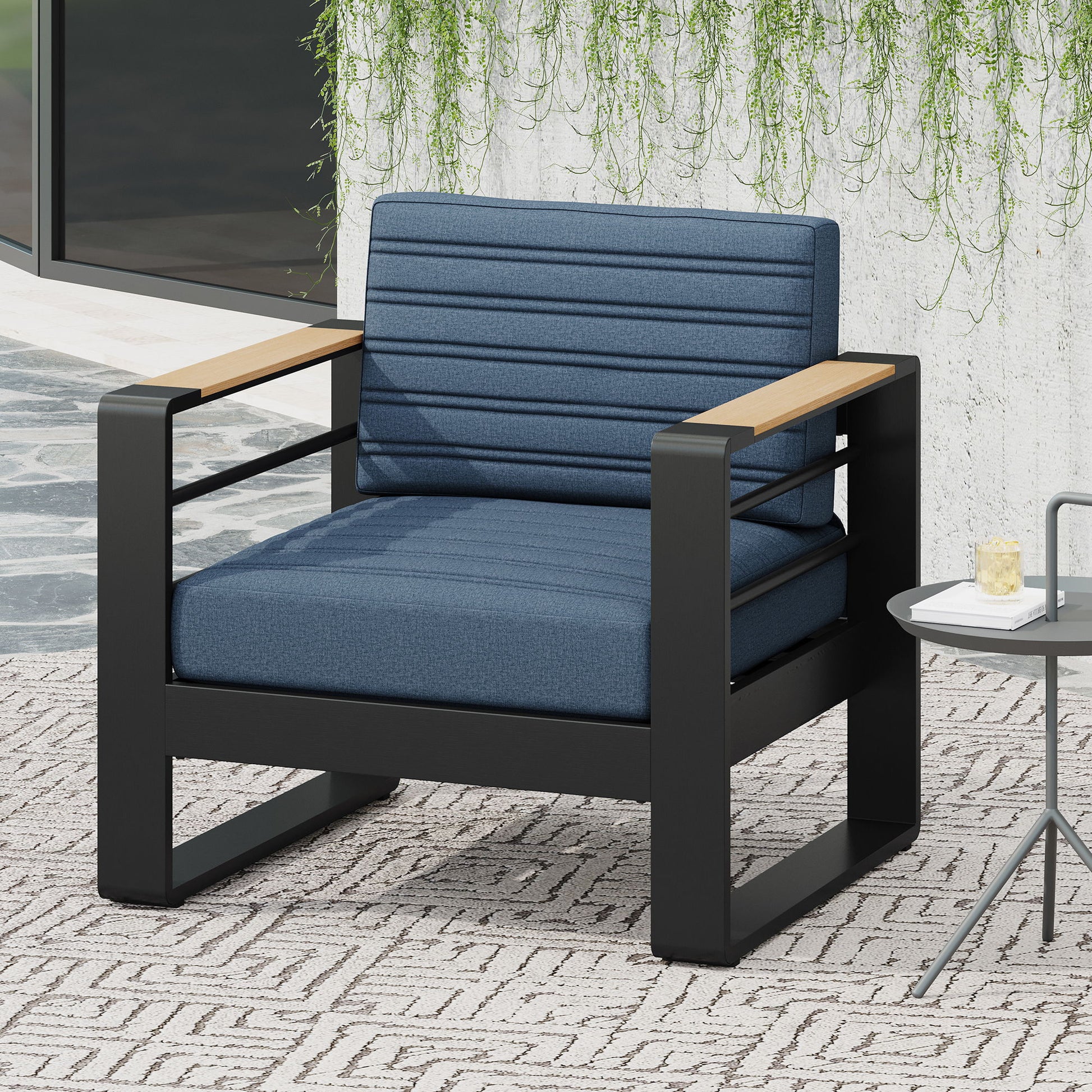 The Giovanna Club Chair Set with an aluminum frame features blue cushions, black metal and light wood armrests. It sits on a patterned rug beside a small round table, with hanging green plants on a concrete wall behind.