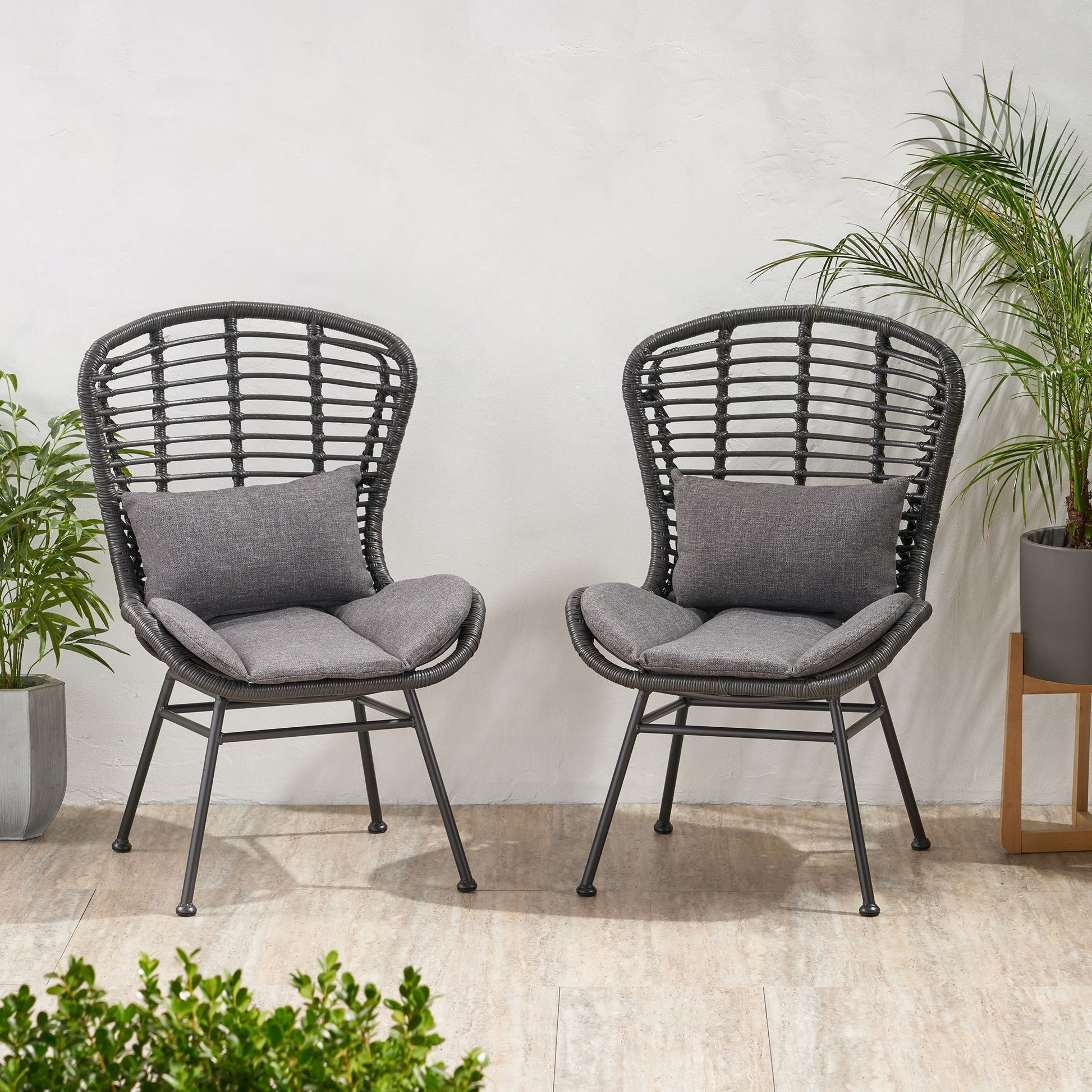 Two Calypso outdoor chairs with water-resistant gray cushions and iron frames sit on a tile floor by a white wall, surrounded by green potted plants for a cozy, inviting setting.