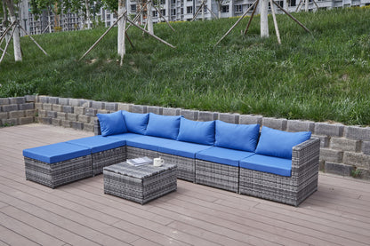 A Patio Sofa With Cushions, featuring blue pillows, sits on a wooden deck beside a low gray stone wall. A matching coffee table is nearby, with a grassy slope and trees in the background.