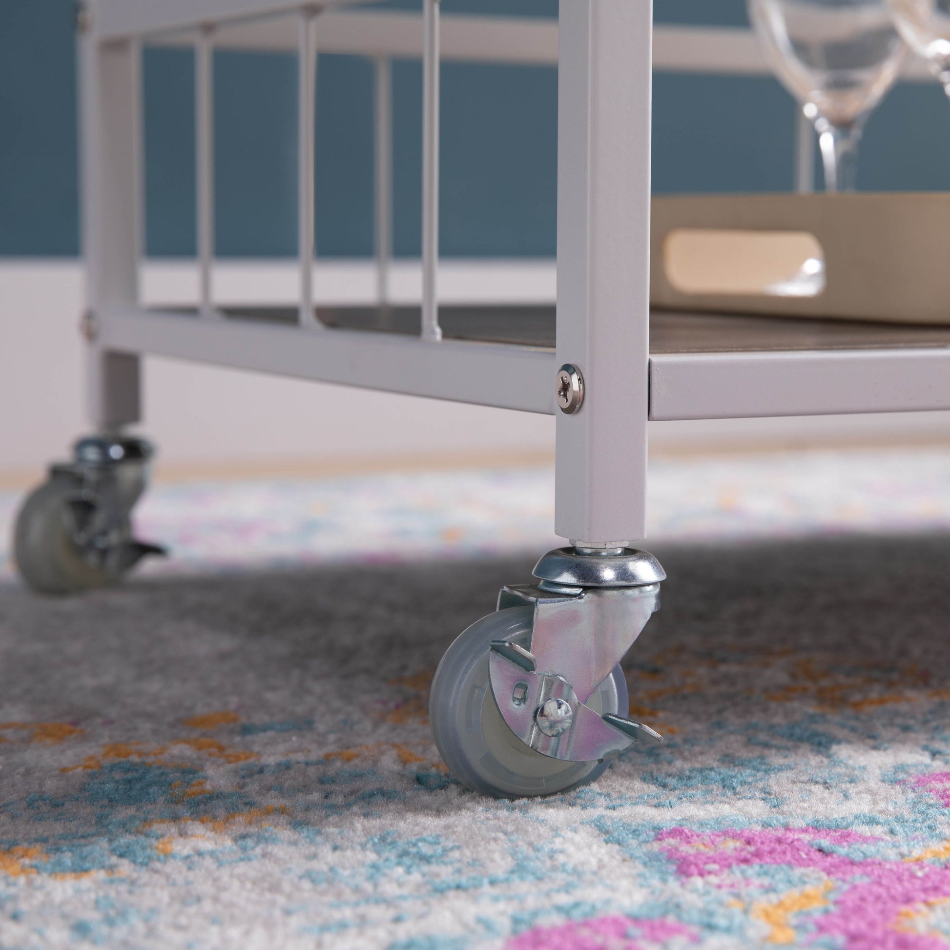 Close-up of the gray caster wheel of the Rolling Kitchen Cart With Storage And Four Wine Bottle Rack on a colorful patterned rug, with a tray and glassware partially visible on the cart above.