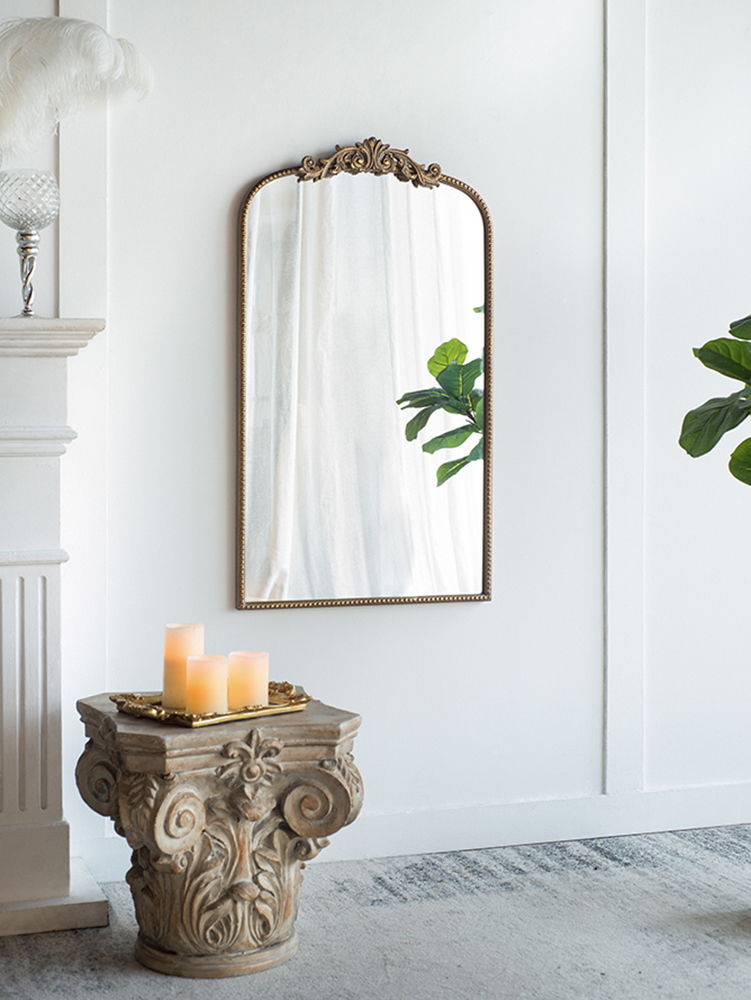 The Arch Mirror, Baroque Inspired Wall Decor, hangs on a white wall. Three lit candles rest on a carved pedestal table in front of it, reflecting green leafy plants and sheer curtains for a serene atmosphere.