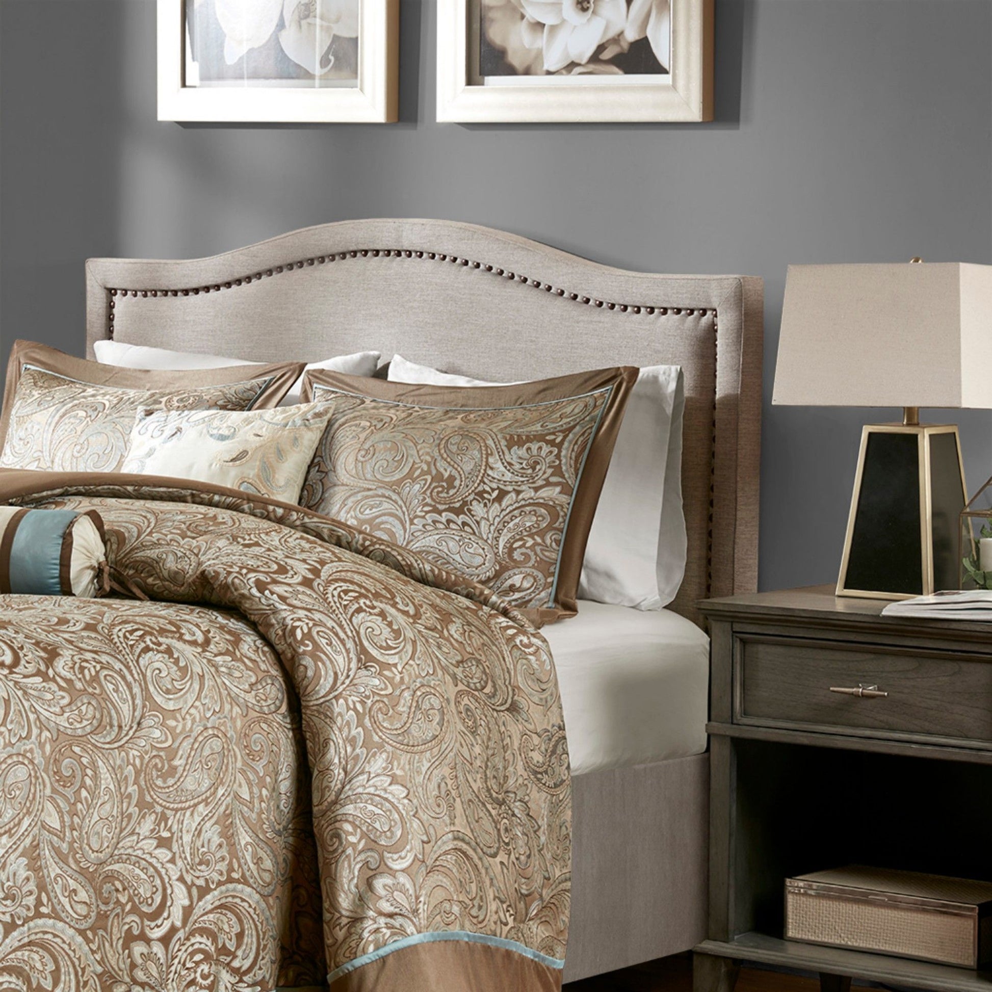 A neatly made bed with a beige and brown paisley comforter, matching pillows, an Elegant Panel Headboard With Copper Nailheads And Adjustable Metal Legs, a wooden nightstand with a modern lamp, and framed floral art on a gray wall.